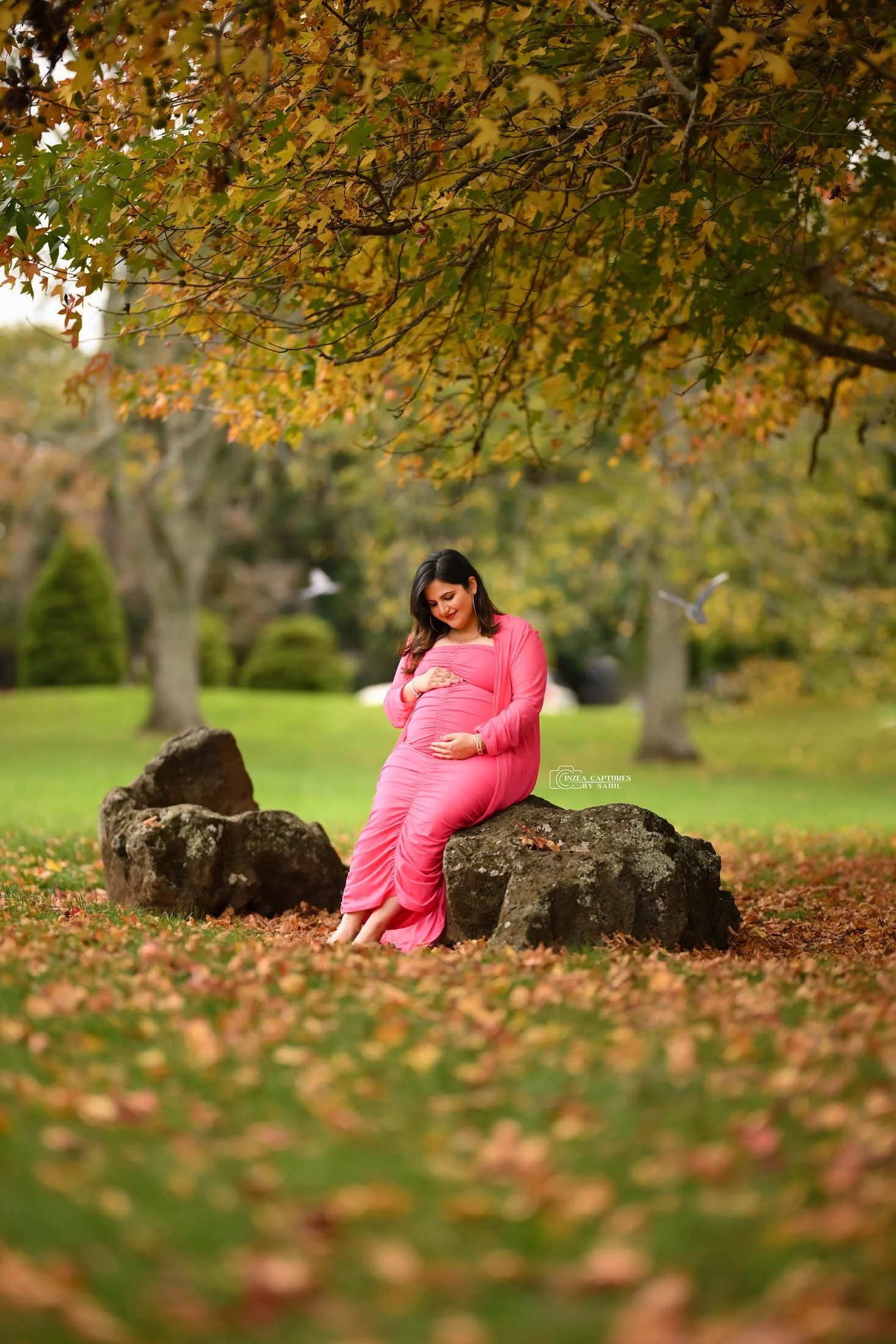 Pregnant woman in pink dress sitting on a rock in a park with fall foliage, looking down at her belly and smiling.