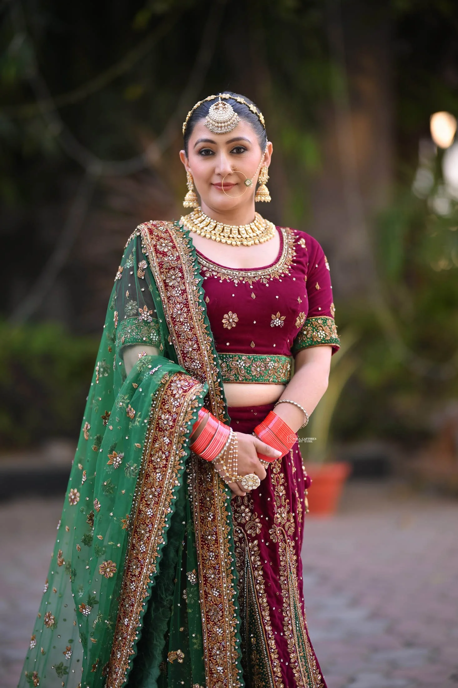 A woman in traditional Indian attire, wearing a maroon lehenga with gold embroidery, gold jewelry, and a green dupatta with detailed embellishments.