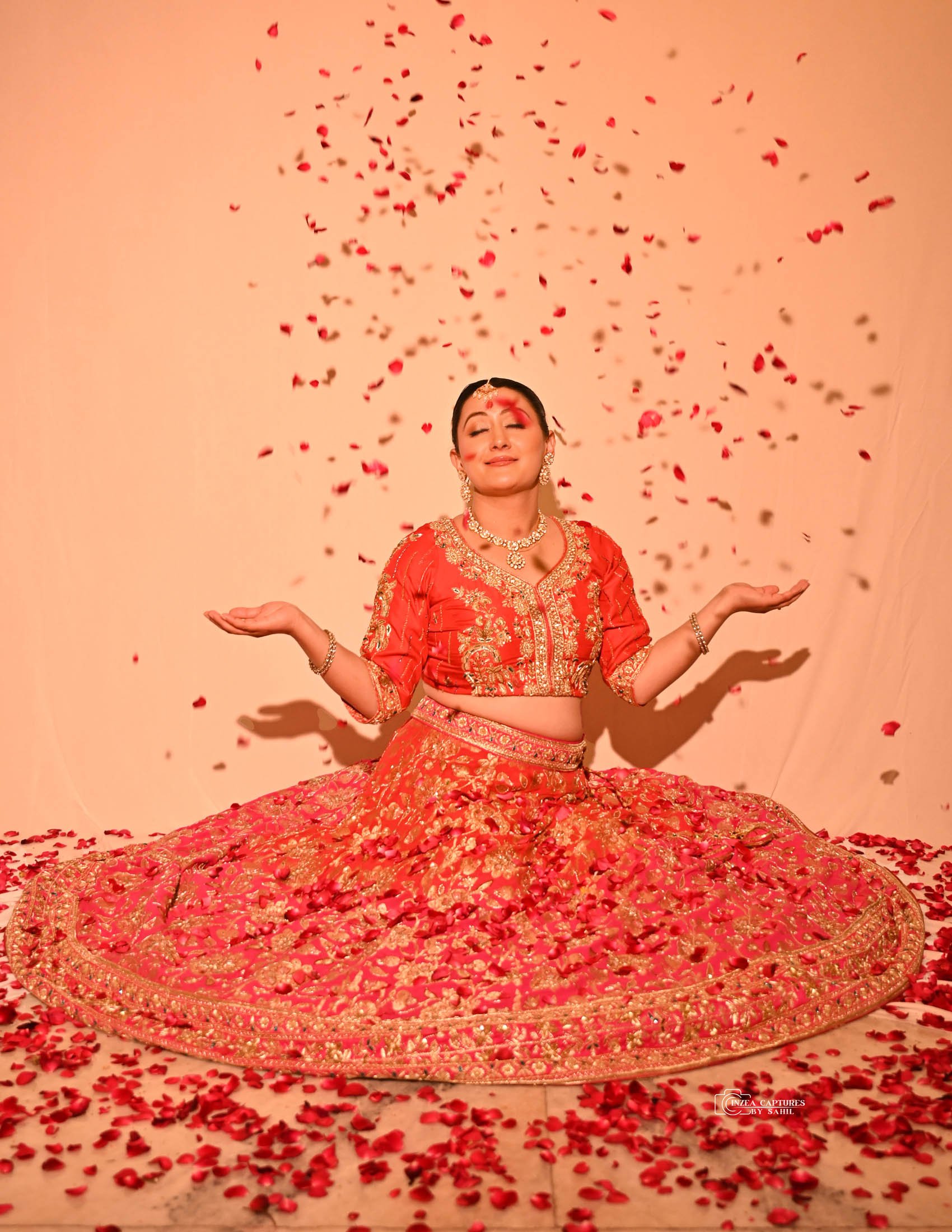 A woman in a red and gold traditional Indian bridal outfit sitting with her arms out, surrounded by falling rose petals, with a content smile.