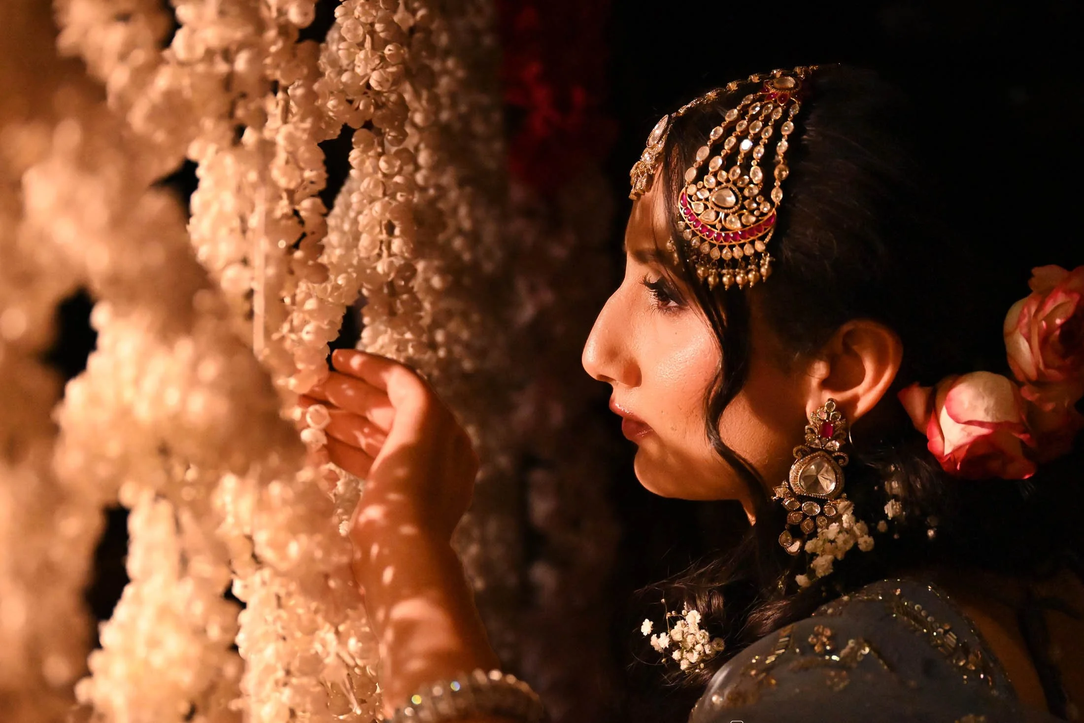 A woman dressed in traditional Indian attire, wearing ornate jewelry, holds and touches hanging strings of white flowers, with pink flowers visible near her ear, in a dimly lit environment.