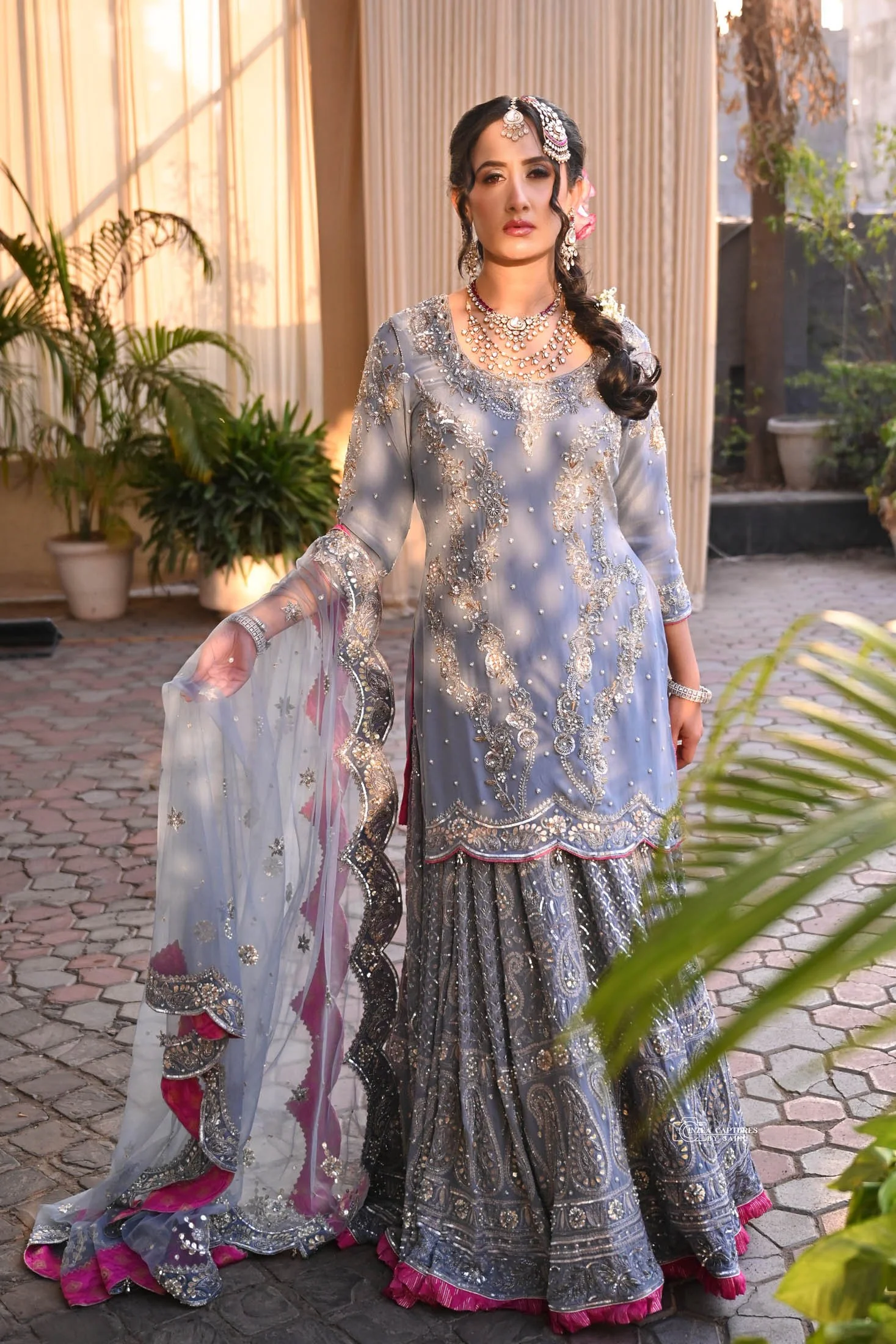 A woman in traditional Pakistani attire standing outdoors in sunlight, wearing a silver-gray embroidered outfit with pink accents, jewelry, and a decorative headpiece.