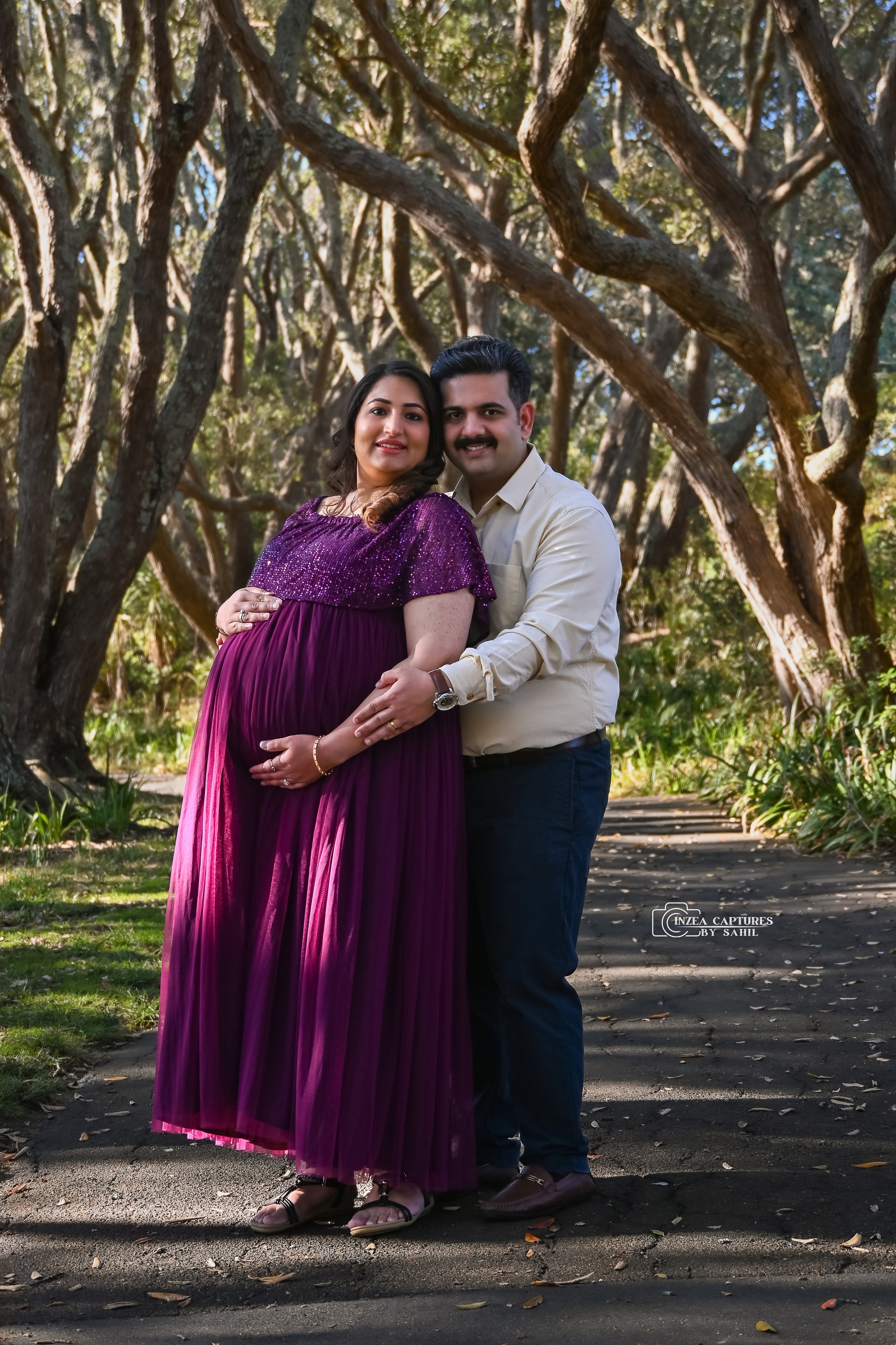 A pregnant woman in a purple dress standing next to a man in a white shirt, both smiling at the camera in a wooded park.
