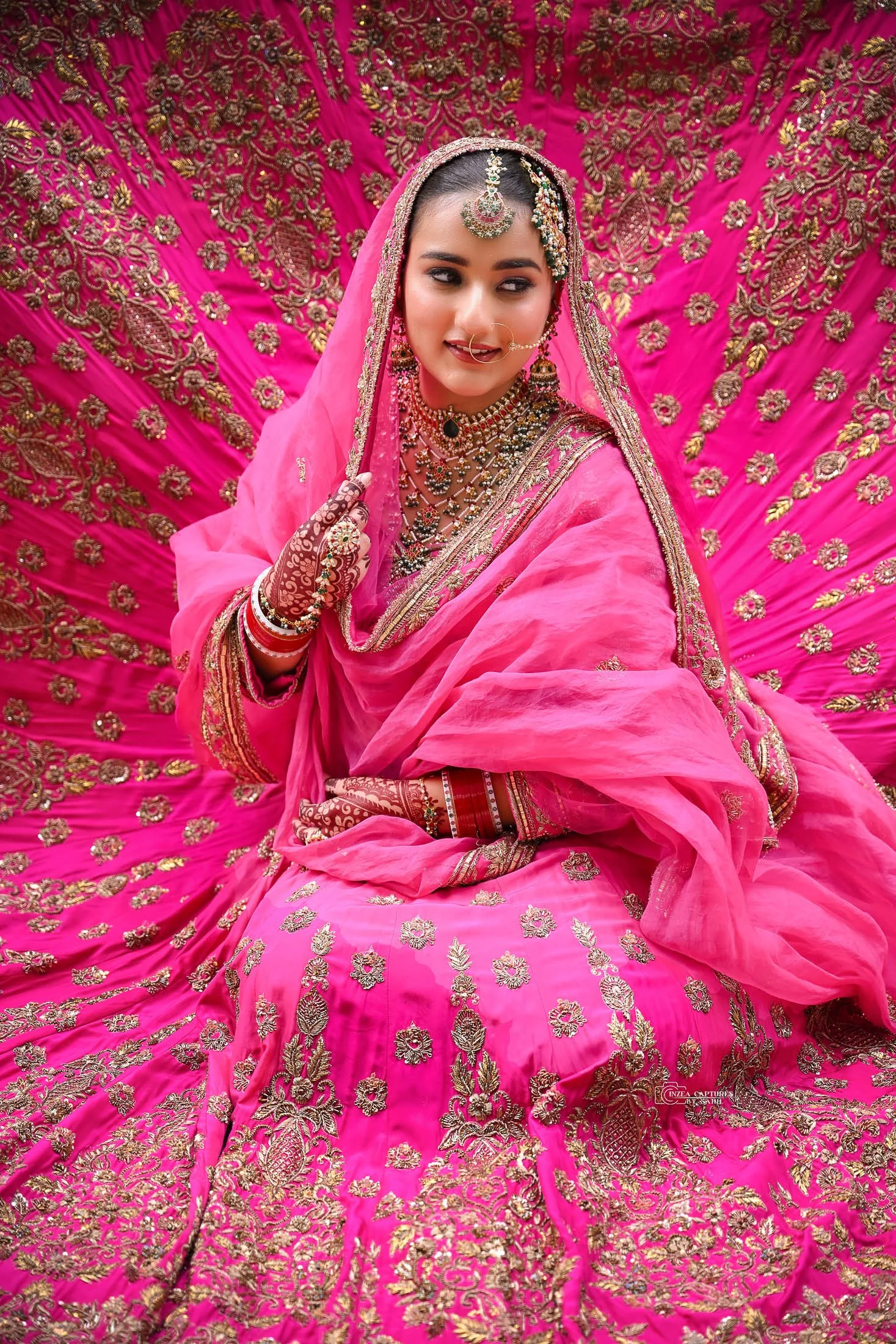 A woman dressed in a vibrant pink traditional Indian bridal outfit, adorned with gold embroidery and jewelry including necklaces, earrings, forehead ornament, and bangles. She is sitting on a matching pink embroidered backdrop.