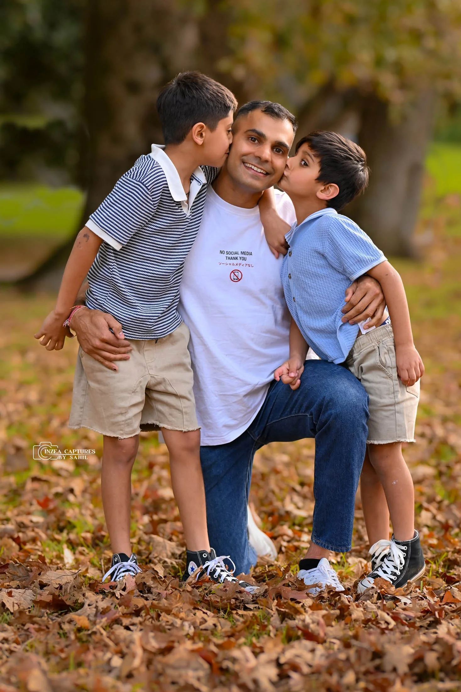 A man kneeling in a park surrounded by fallen autumn leaves, with two young boys giving him kisses on each cheek. The man is smiling and wearing a white T-shirt and jeans, while the boys are wearing casual shirts, shorts, and sneakers.