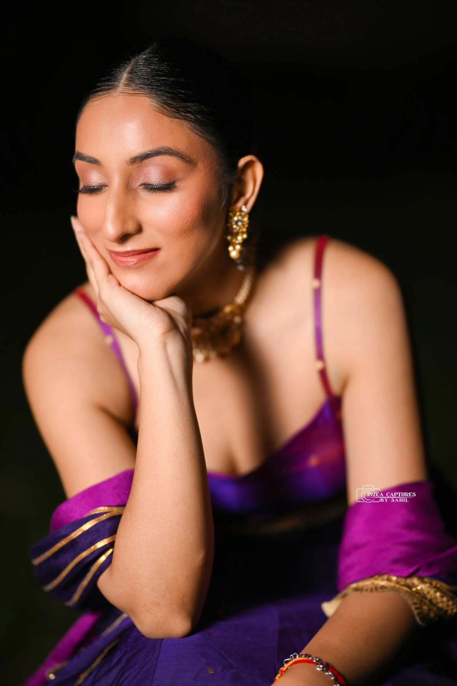 A woman with closed eyes, wearing traditional Indian attire, gold jewelry, and a purple and pink saree, resting her chin on her hand.