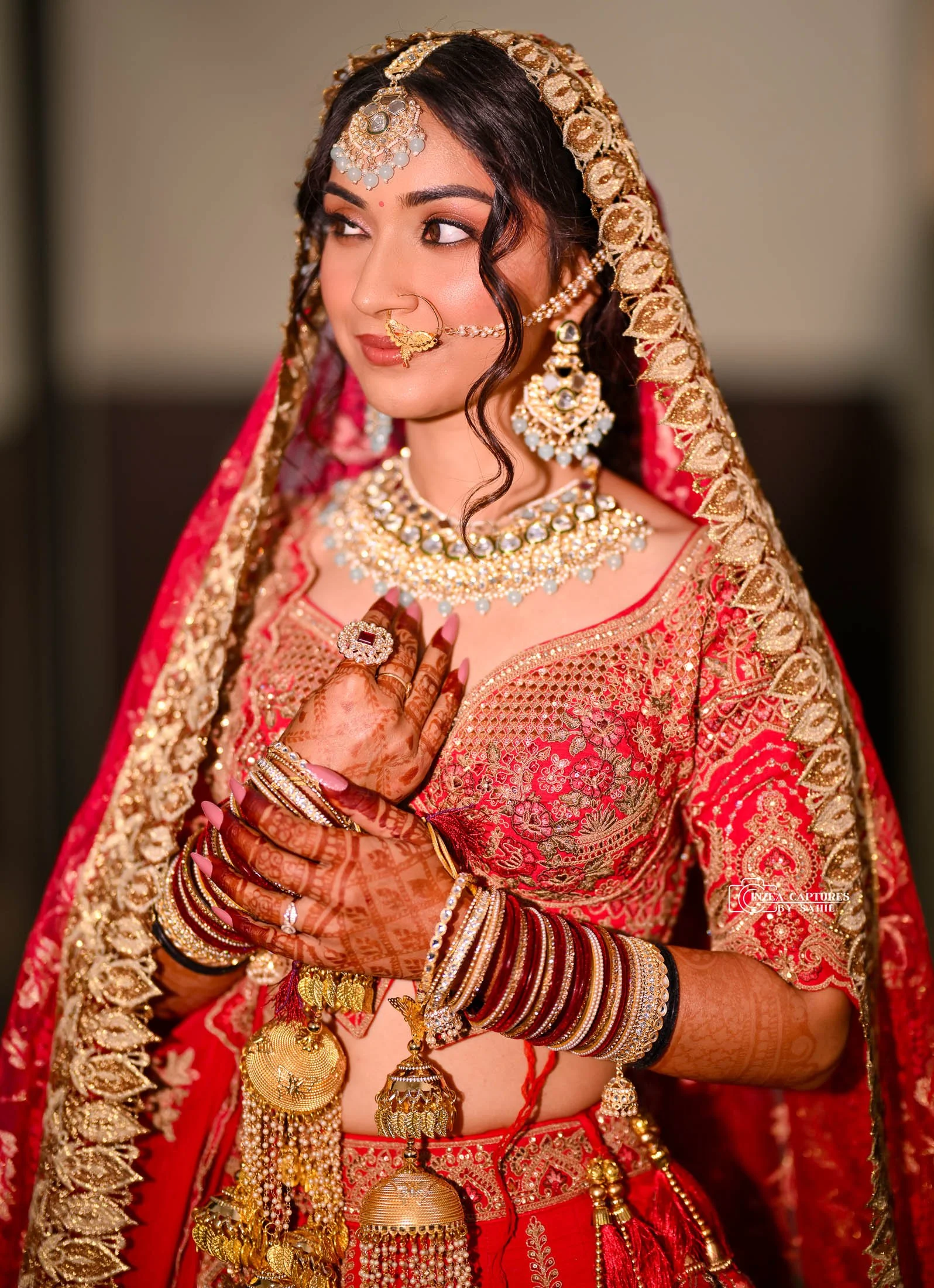 A woman dressed in traditional Indian bridal attire, wearing a red and gold saree, ornate jewelry including necklaces, earrings, a nose ring, and bangles, with henna designs on her hands, posing with her hands crossed over her chest.