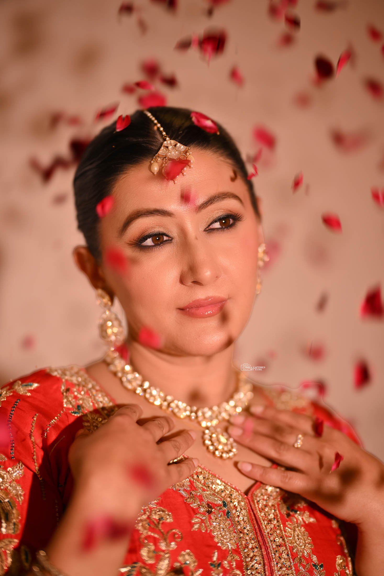 A woman dressed in traditional Indian attire, with jewelry and henna on her hands, is surrounded by falling rose petals.