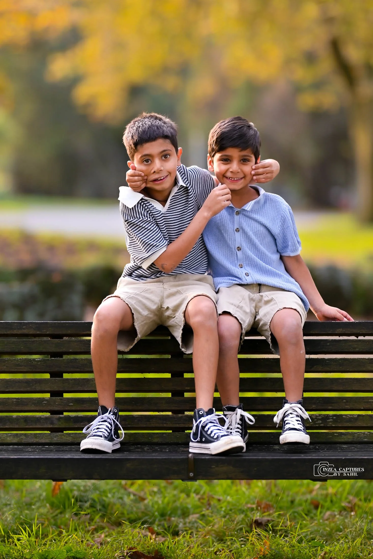 Two young boys sitting on a park bench, smiling and playing happily. One boy has his arm around the other, and both are dressed in casual shorts and sneakers. The background shows blurred trees and autumn scenery.