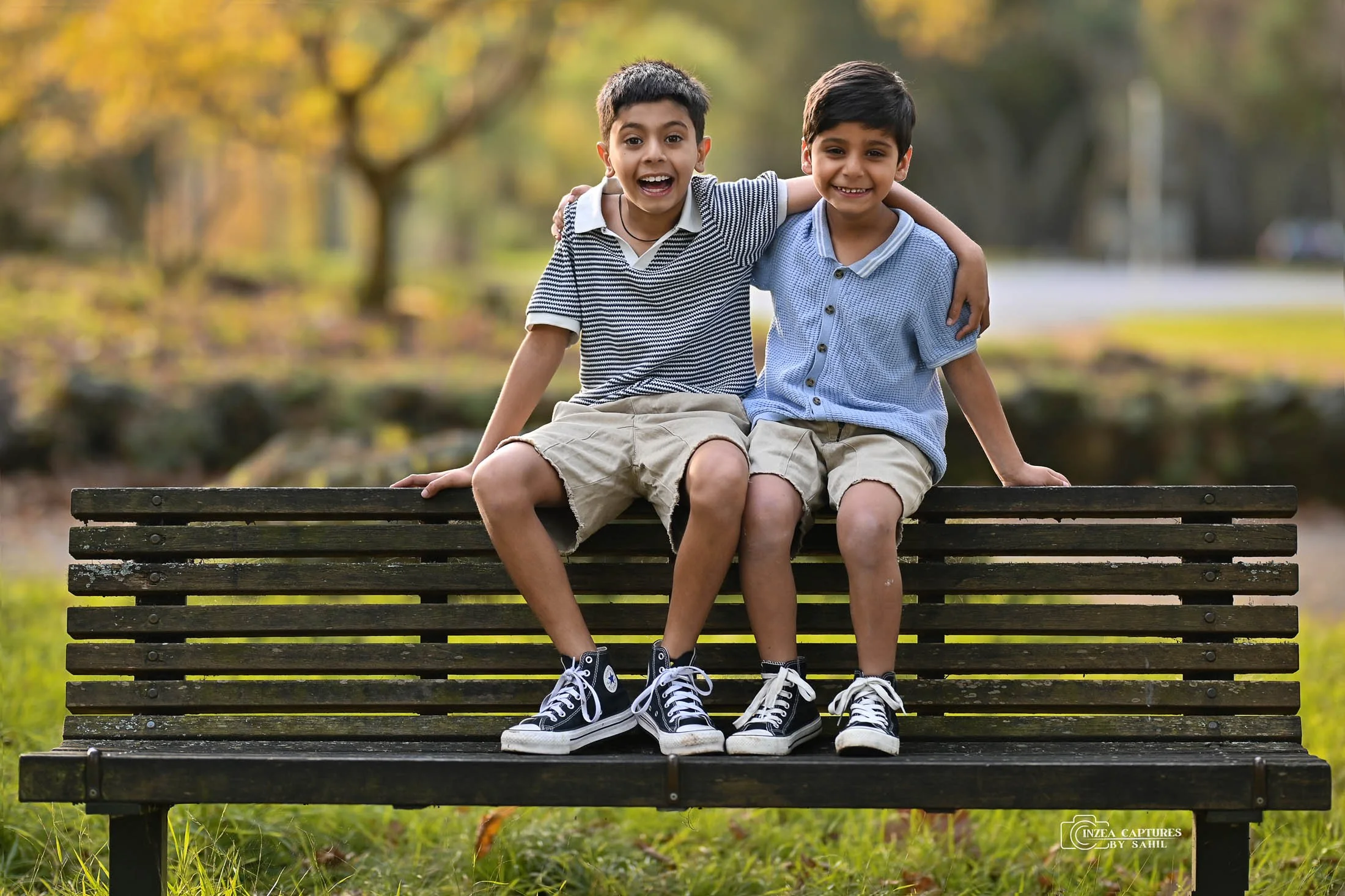 Two young boys sitting on a park bench with their arms around each other, smiling. They are dressed in casual summer clothes, with sneakers, in a park with trees and grass.