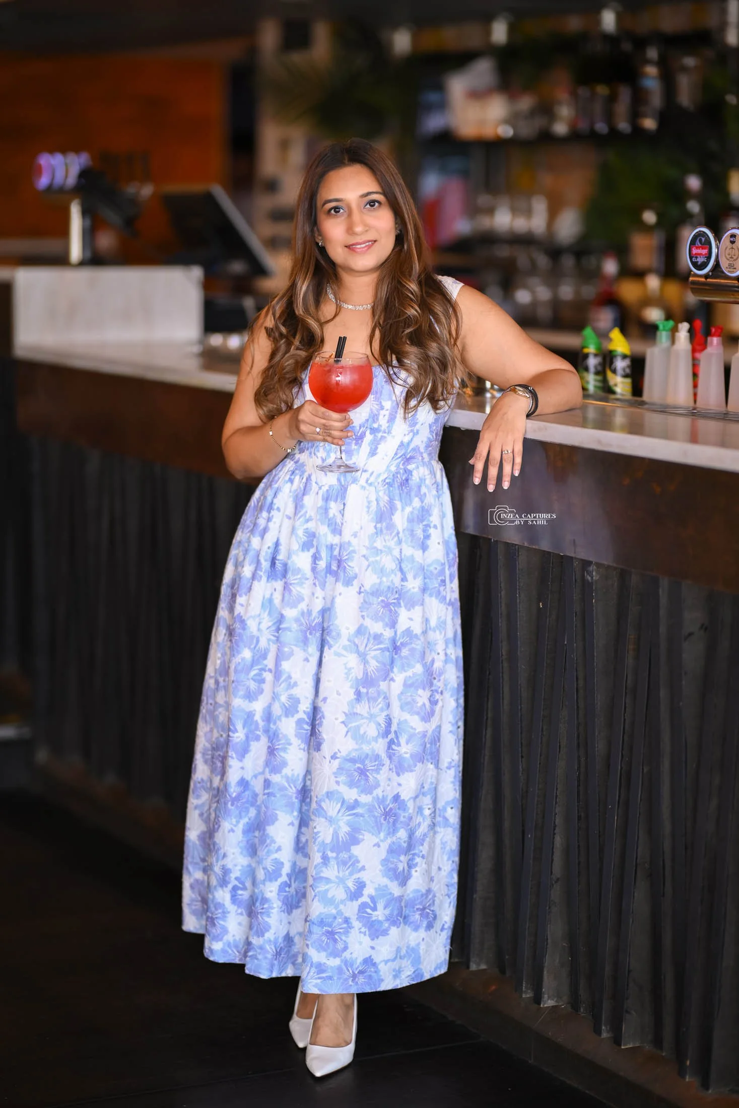 A woman in a blue and white floral dress stands at a bar, holding a red cocktail glass with a straw, smiling at the camera.