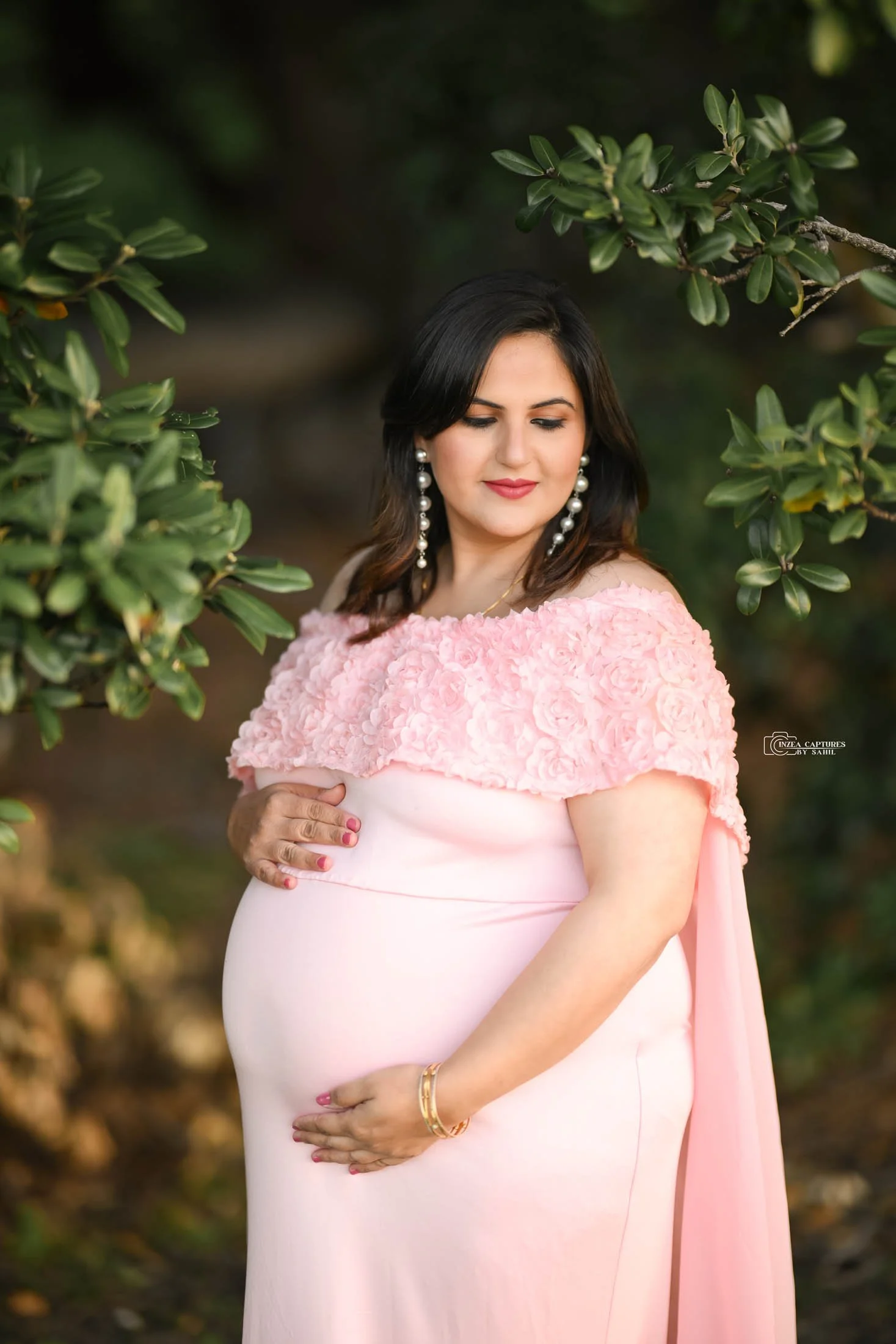 A pregnant woman wearing a pink dress with a floral textured top, standing outdoors among greenery, gently holding her baby bump, with earrings and a gold bracelet.