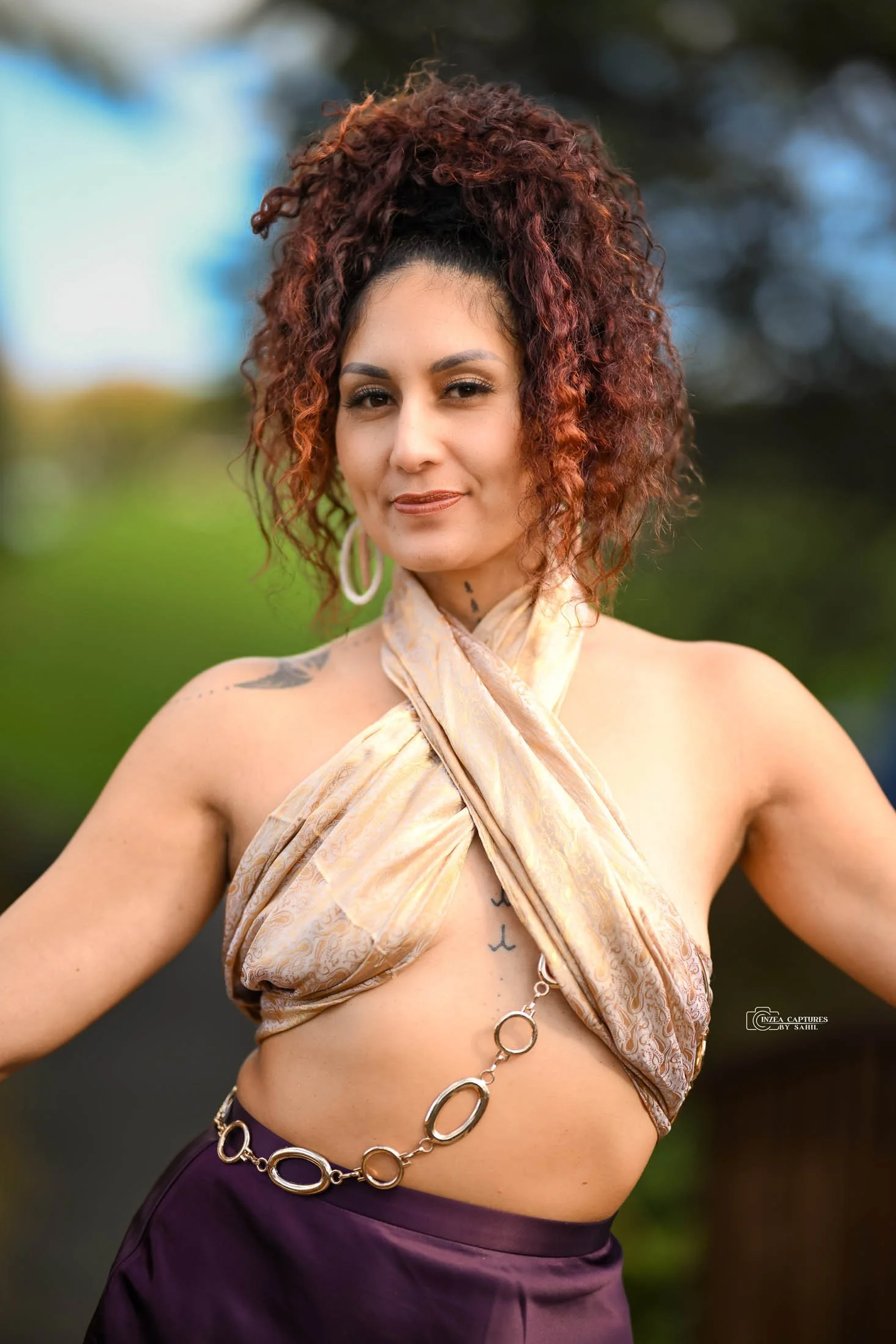 A woman with curly auburn hair, wearing a beige silk halter top with a golden chain belt, standing outdoors with a blurred green background.