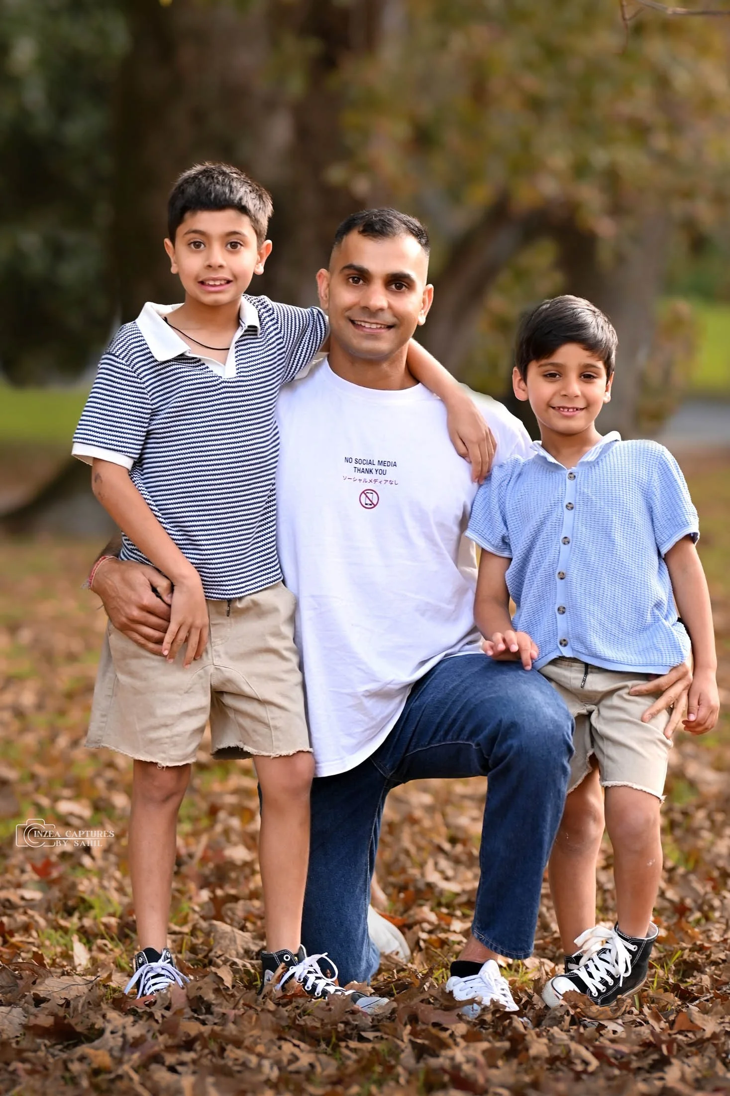 A man kneeling outdoors with two young boys, all smiling, surrounded by fallen autumn leaves with trees in the background.