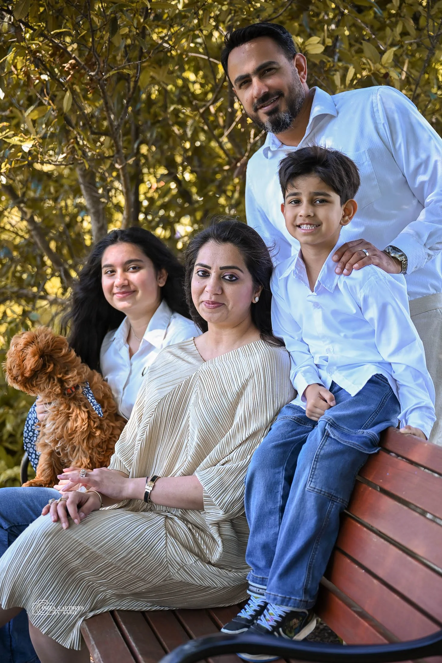 Family portrait of five people and a dog sitting outdoors on a park bench, surrounded by green foliage, smiling at the camera.