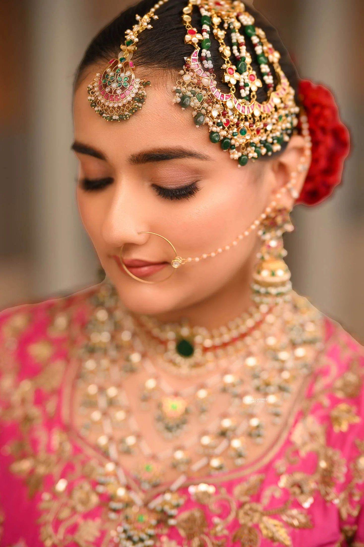 A woman dressed in traditional Indian bridal attire, wearing an elaborate pink and gold embroidered outfit. She has intricate jewelry including a large headpiece, earrings, a nose ring connected with a chain, and layered necklaces. Her eyes are close