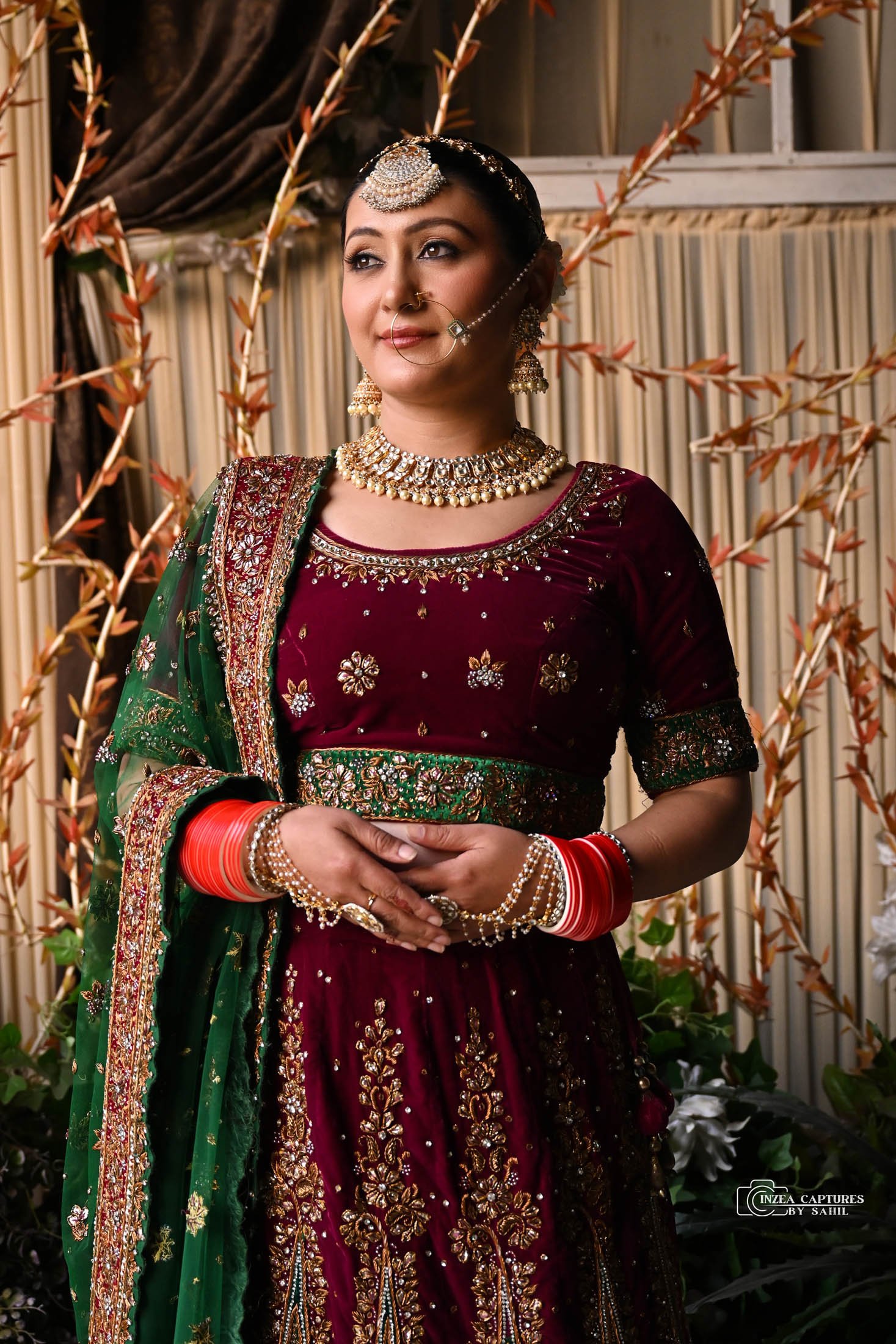 A woman dressed in traditional Indian bridal attire, wearing a rich maroon and green embroidered lehenga, with heavy gold jewelry and red bangles. She has a nose ring and decorative headpiece, standing amidst decorative background with plants and dra