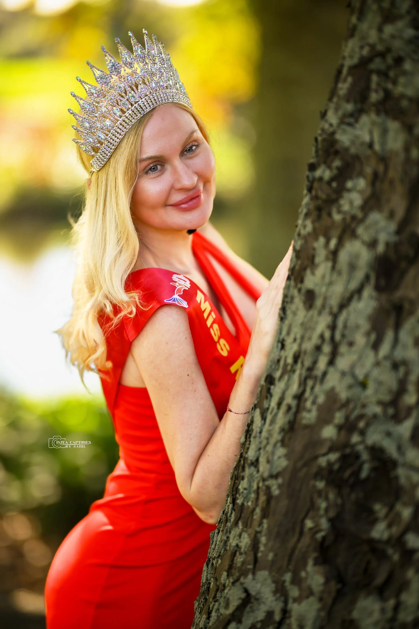 A woman in a red dress and a crown, holding a tree trunk outdoors with a blurred background of foliage and sunlight.