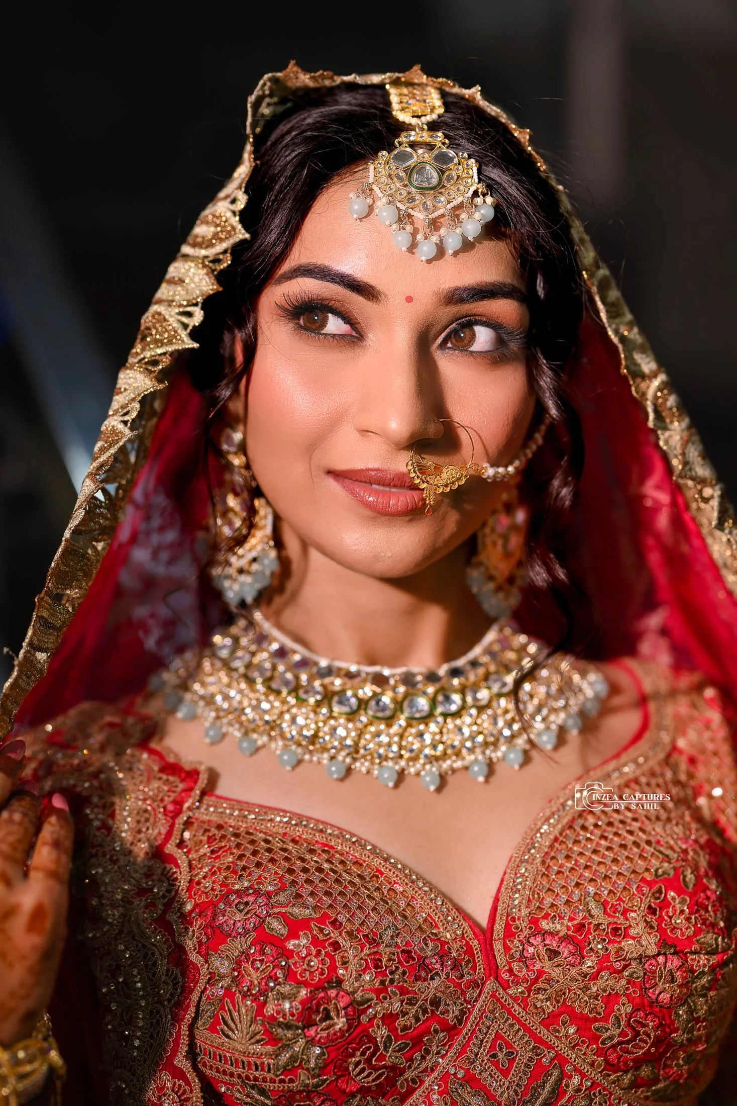 A woman dressed in traditional Indian bridal attire, wearing red and gold embroidery, with gold and pearl jewelry, a nose ring, and a head covering with gold accents.
