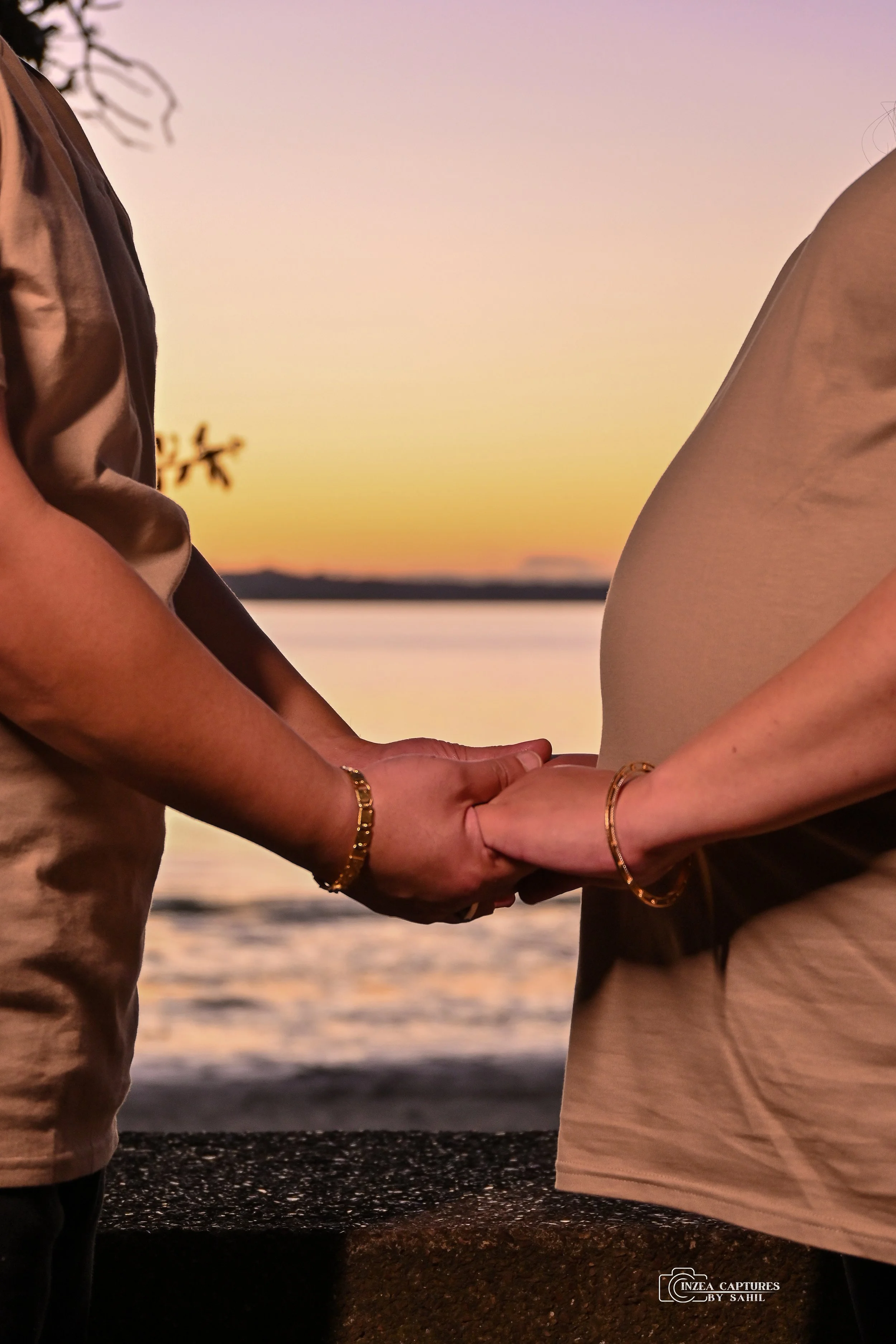 A couple holding hands during sunset by a body of water, with trees and hills in the background.