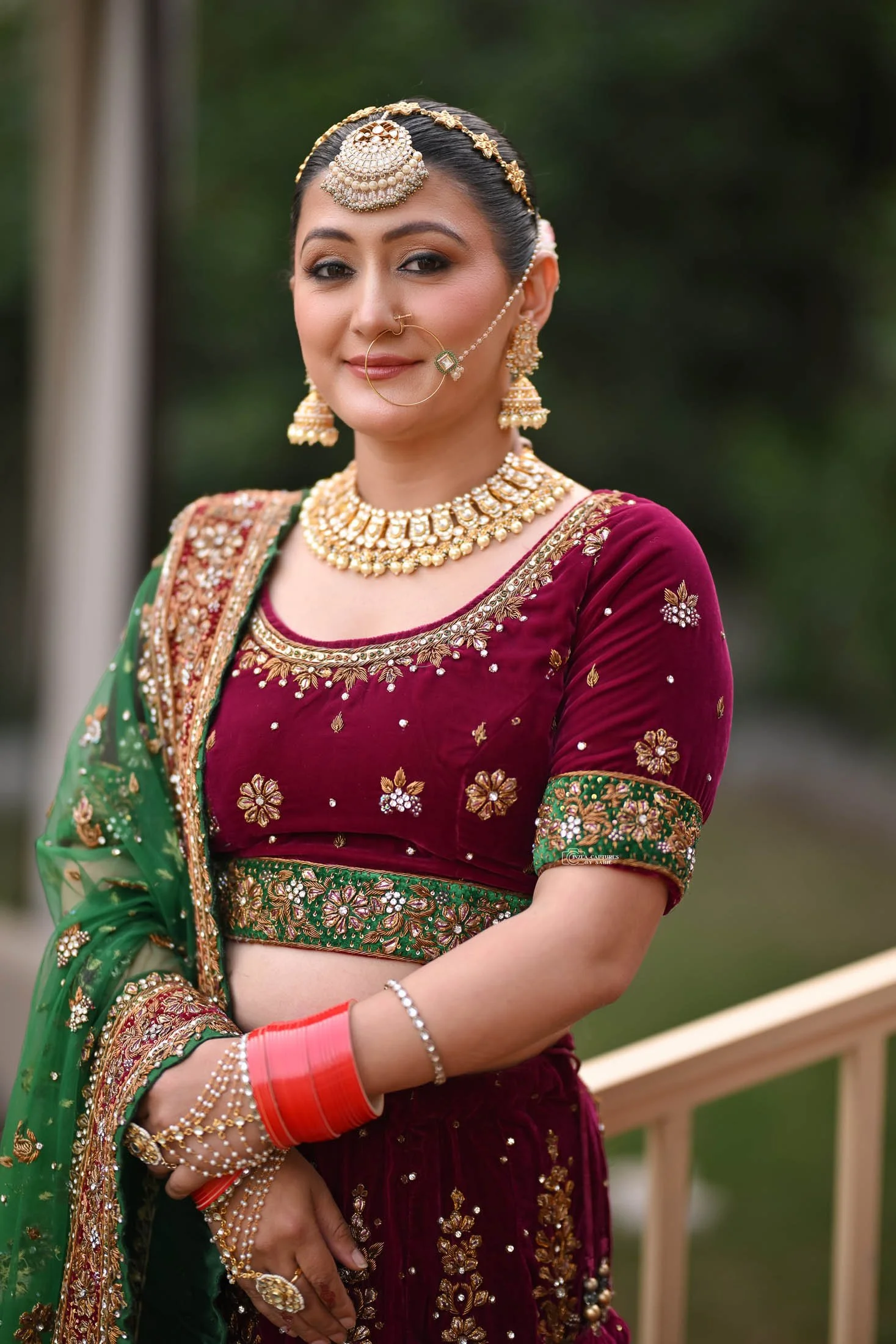 A woman dressed in traditional Indian attire poses outdoors, wearing a maroon velvet outfit with intricate gold embroidery, and adorned with elaborate gold jewelry including a headpiece, earrings, necklace, nose ring, bangles, and rings.