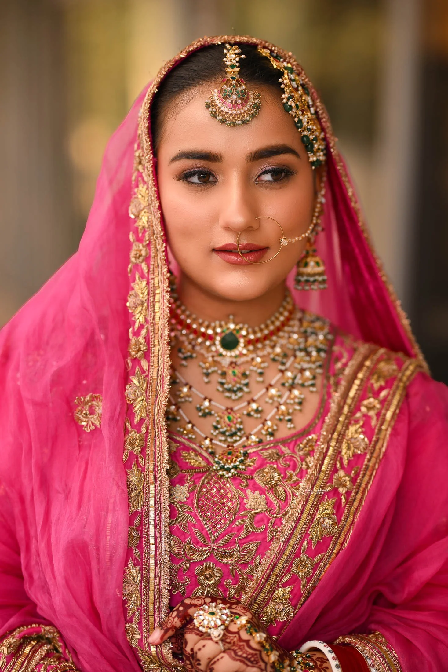 A woman dressed in traditional Indian bridal attire with vibrant pink and gold embroidered clothing, intricate jewelry, and henna on her hand.
