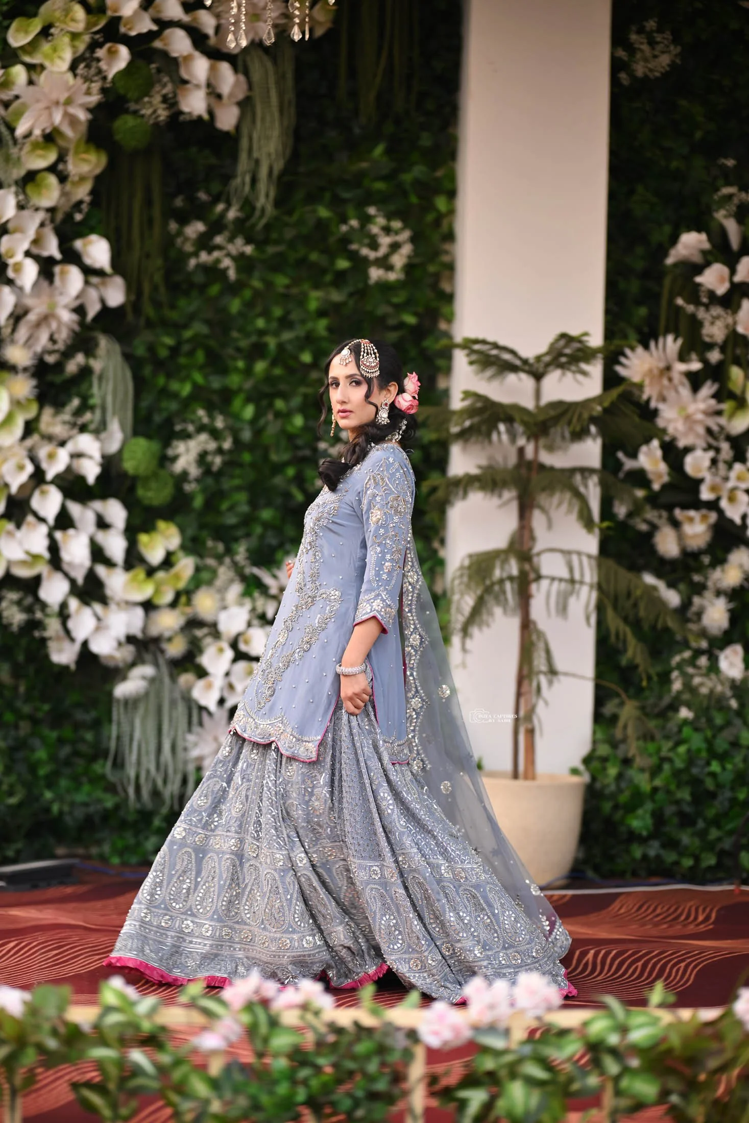 A woman in traditional South Asian attire at a formal event, standing on a decorated stage with floral arrangements and green foliage in the background.