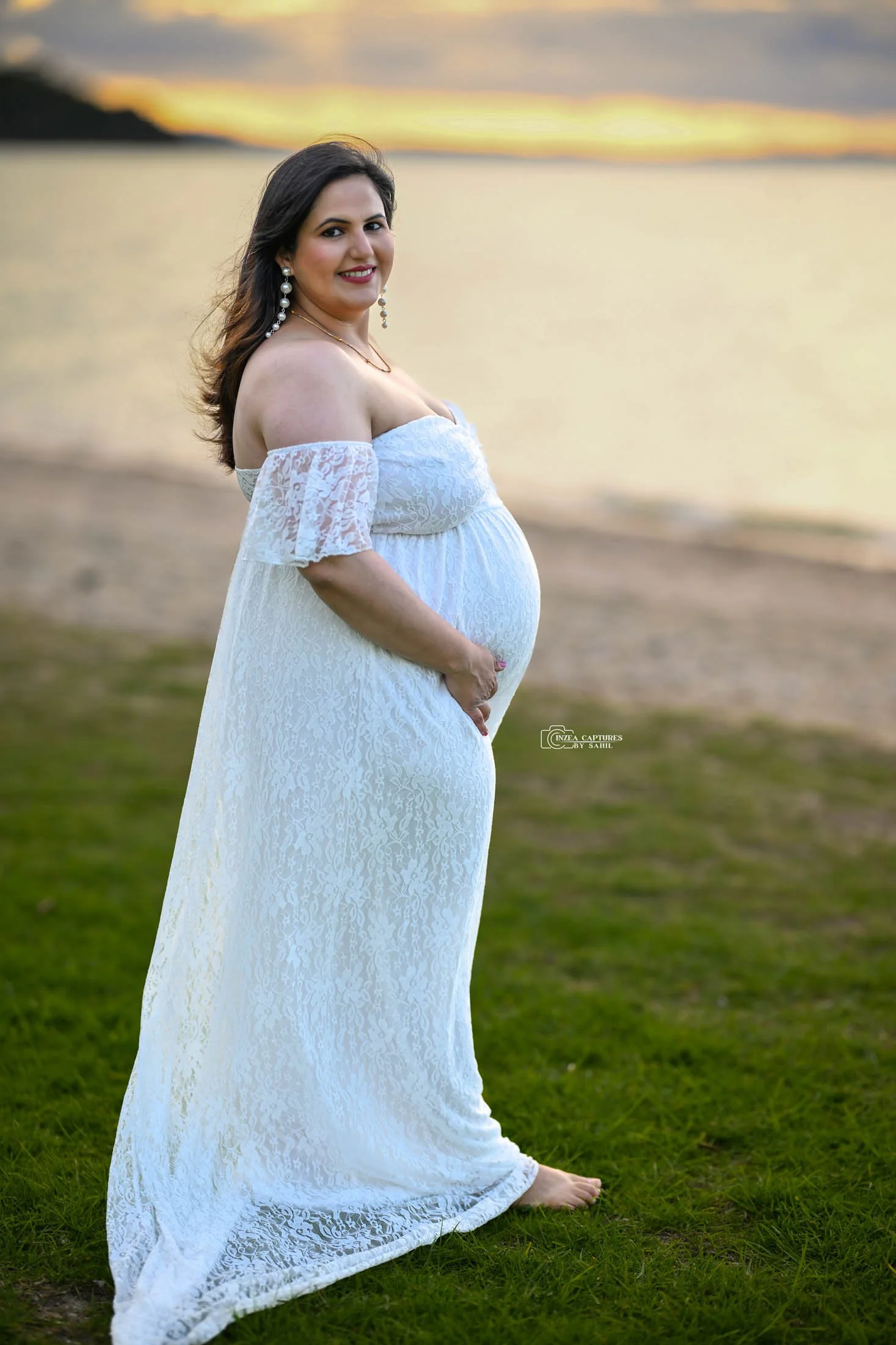 Pregnant woman in a white lace off-shoulder dress standing on grass near a body of water at sunset.