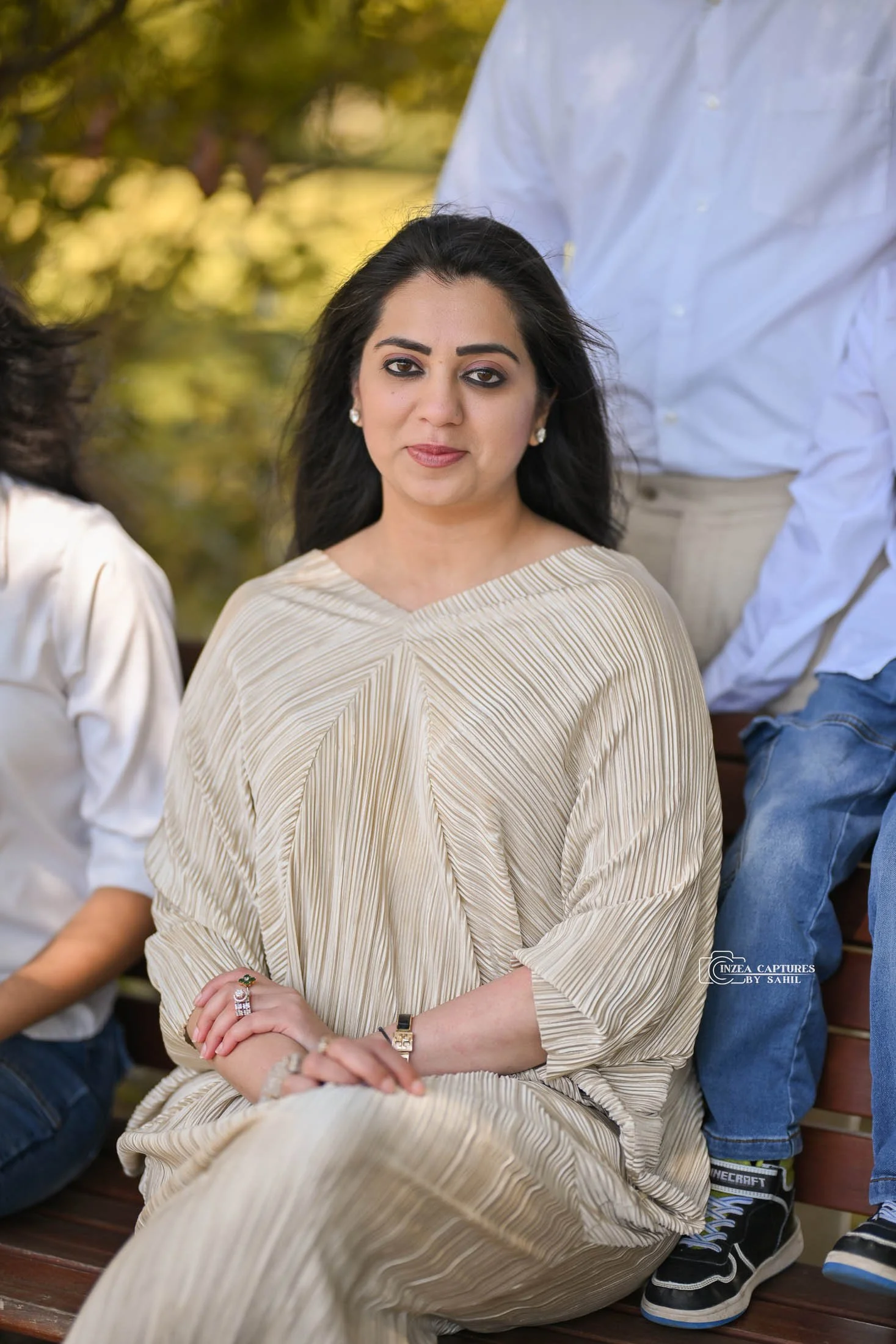 A woman with dark hair and eyes sitting on a park bench, wearing a beige pleated dress, with a small group of people around her.