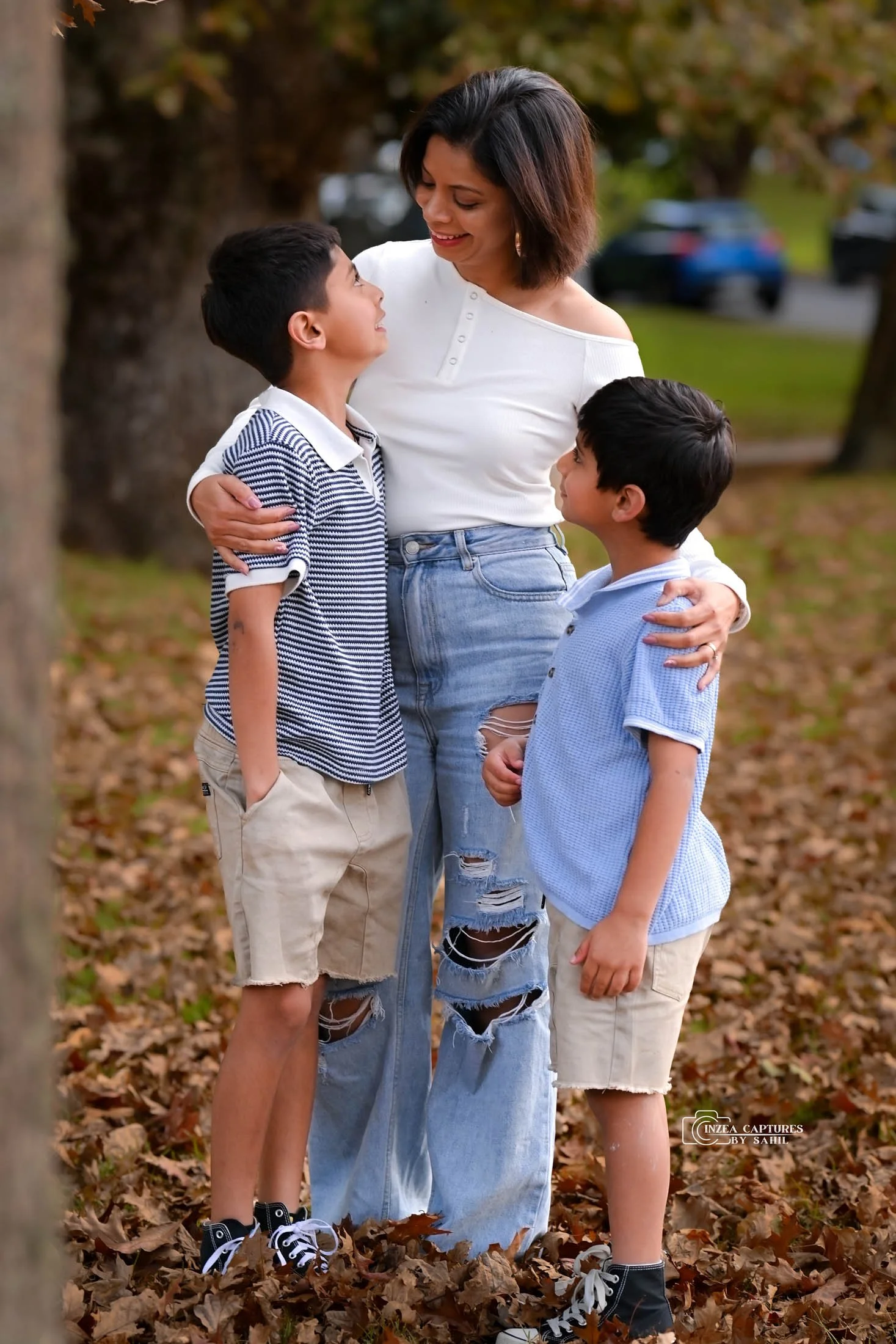 A woman with short dark hair and wearing ripped jeans is hugging and smiling at two young boys outdoors in a park, surrounded by fallen leaves.
