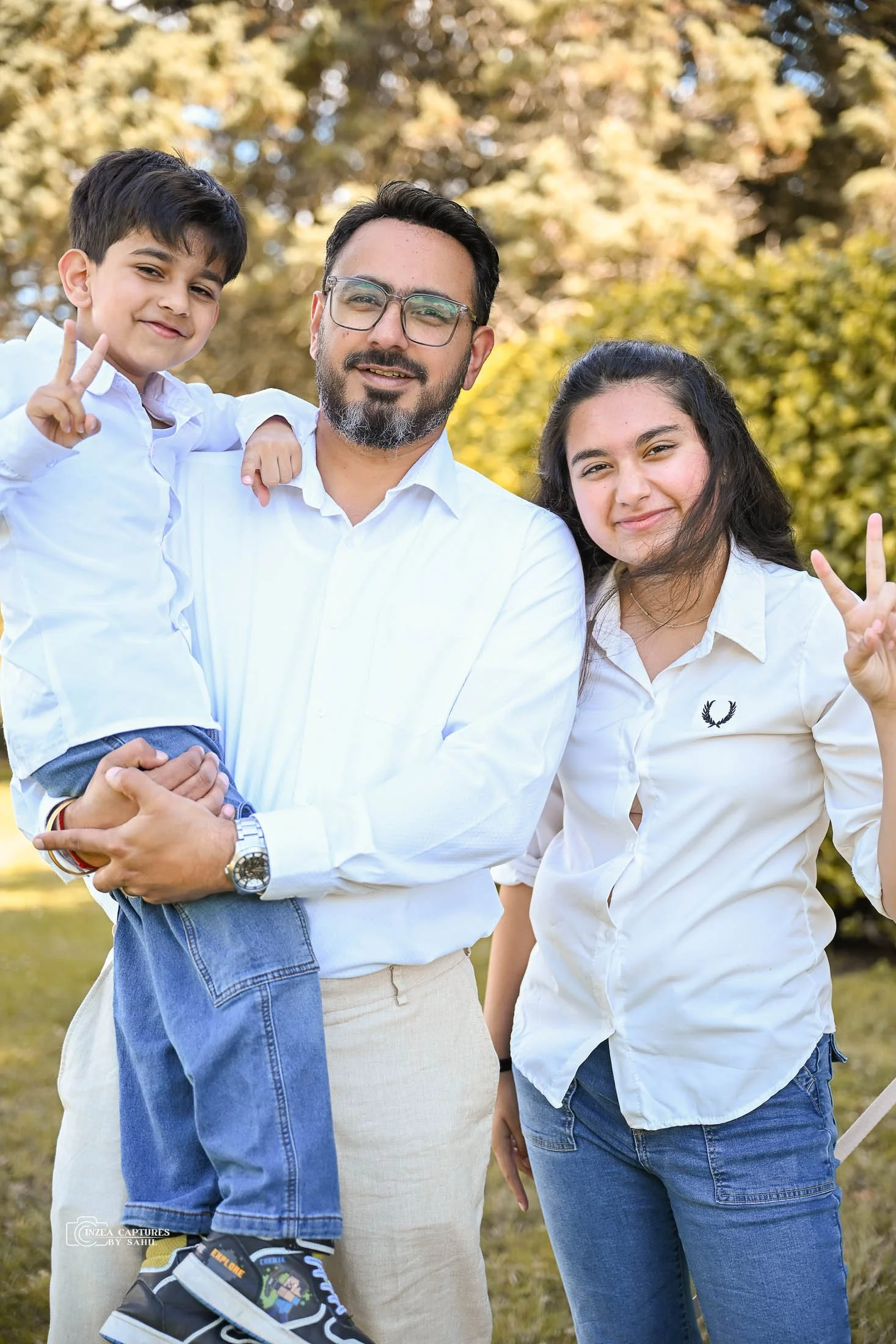 A man holding a young boy in a white shirt and jeans, with a woman standing beside them, all outdoors with trees in the background.