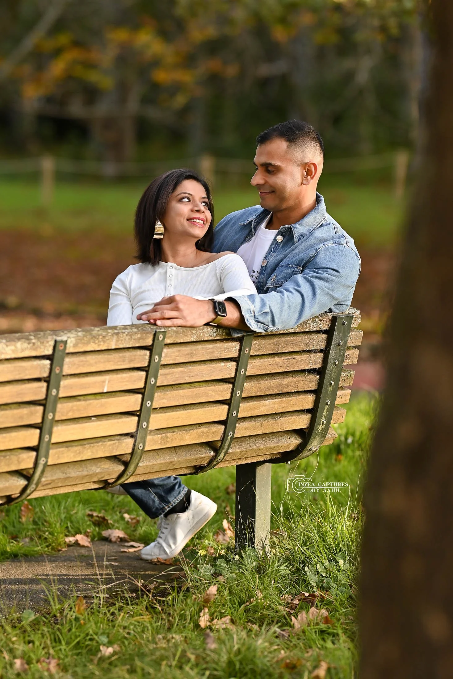 A couple sitting on a park bench outdoors, looking at each other and smiling, surrounded by trees and fallen leaves.