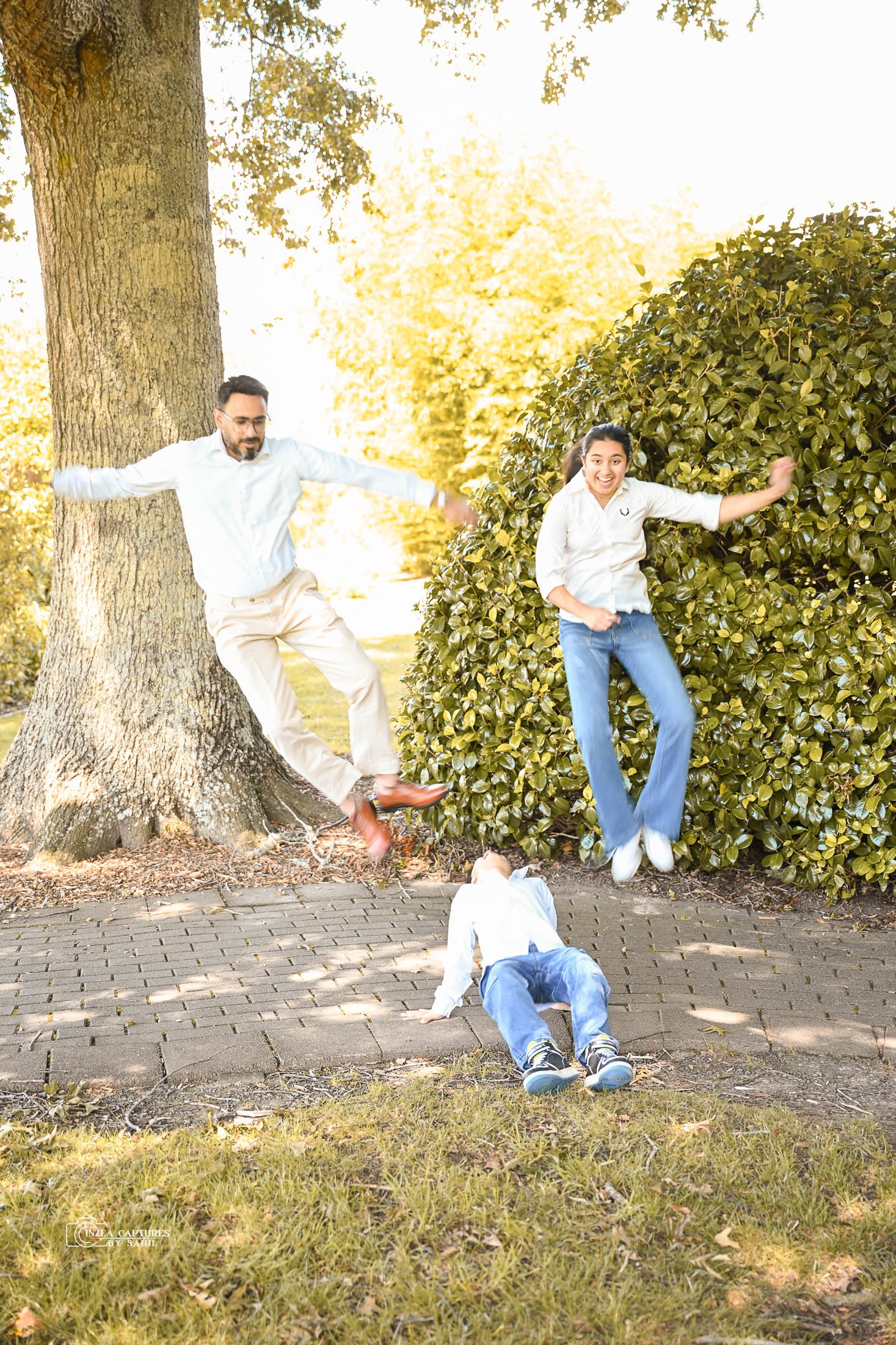 Family of three having fun outdoors on a sunny day, with parents jumping and child on the ground near a tree and bush.