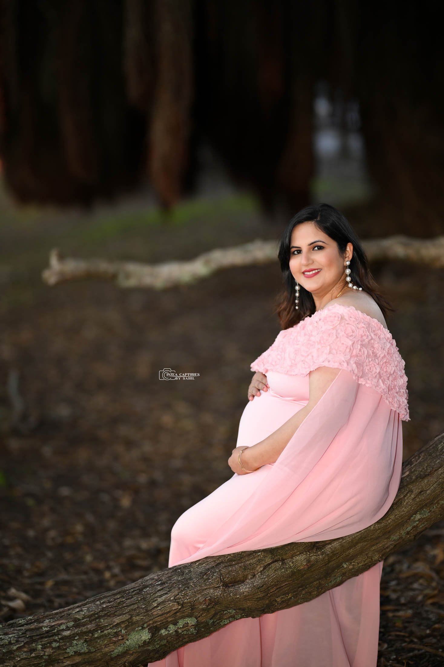 A pregnant woman in a pink dress sitting on a tree branch outdoors.