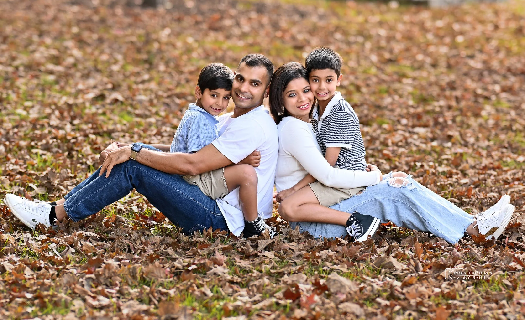 A happy family of four sitting back-to-back on a bed of fallen autumn leaves in a park, smiling at the camera.