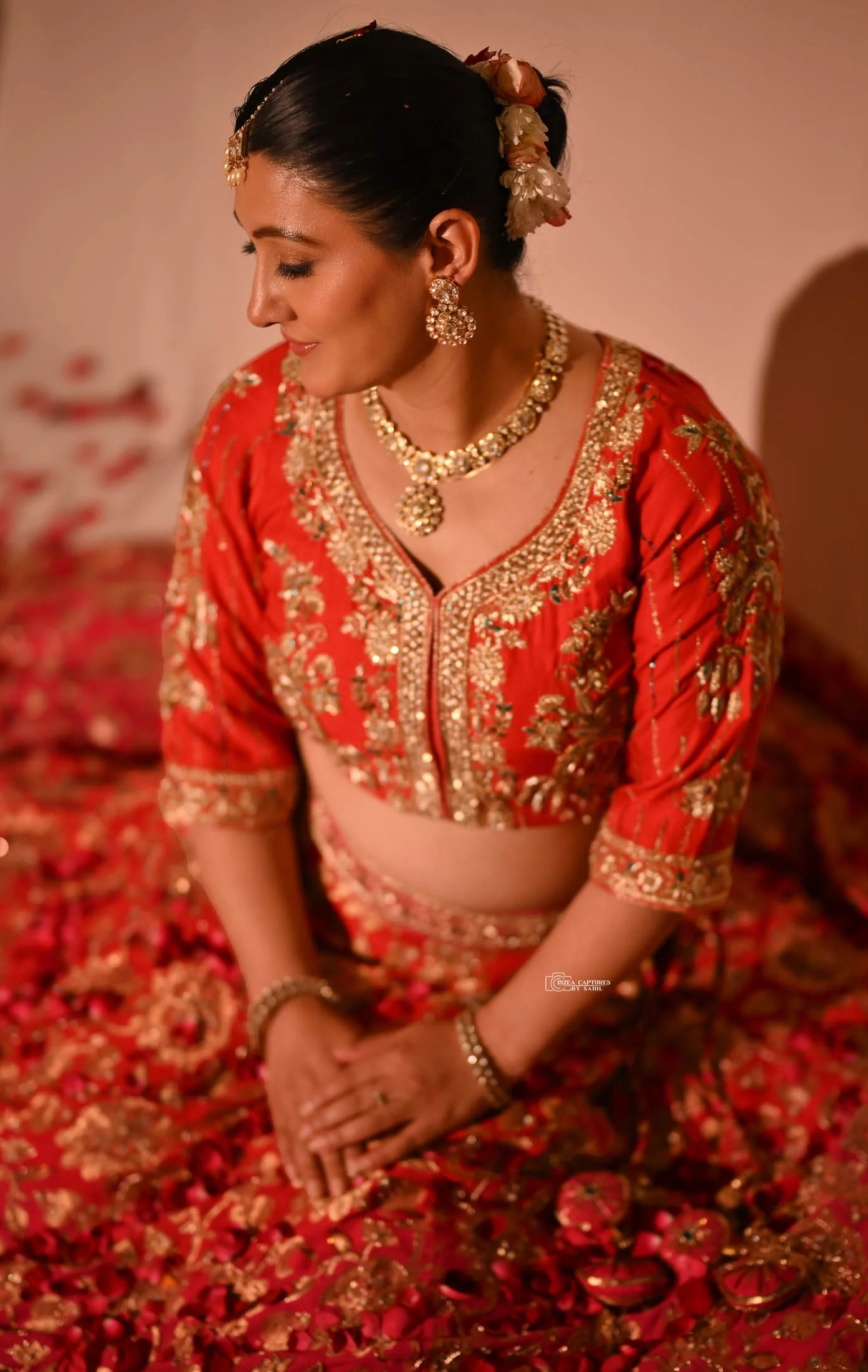 A woman dressed in traditional red and gold embroidered attire, sitting with her eyes closed amidst scattered rose petals. She is wearing jewelry including earrings, a necklace, and bracelets, with flowers adorning her hair.