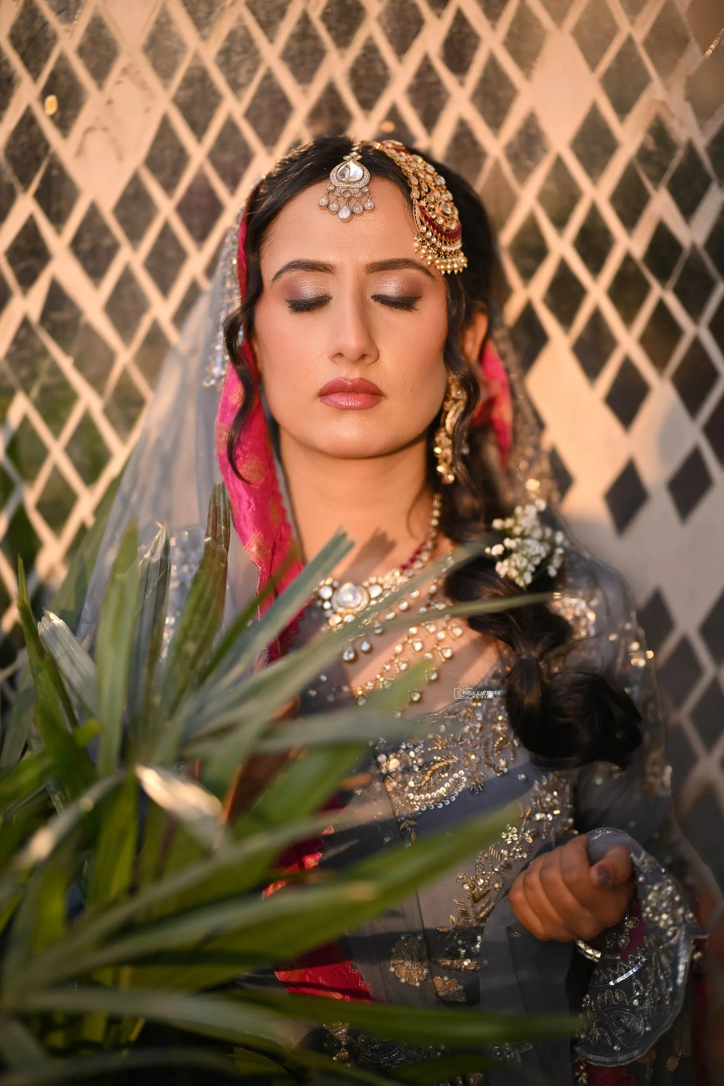 A woman in traditional Indian attire with elaborate jewelry and makeup, posed with her eyes closed in front of a lattice-patterned background, holding a pineapple.