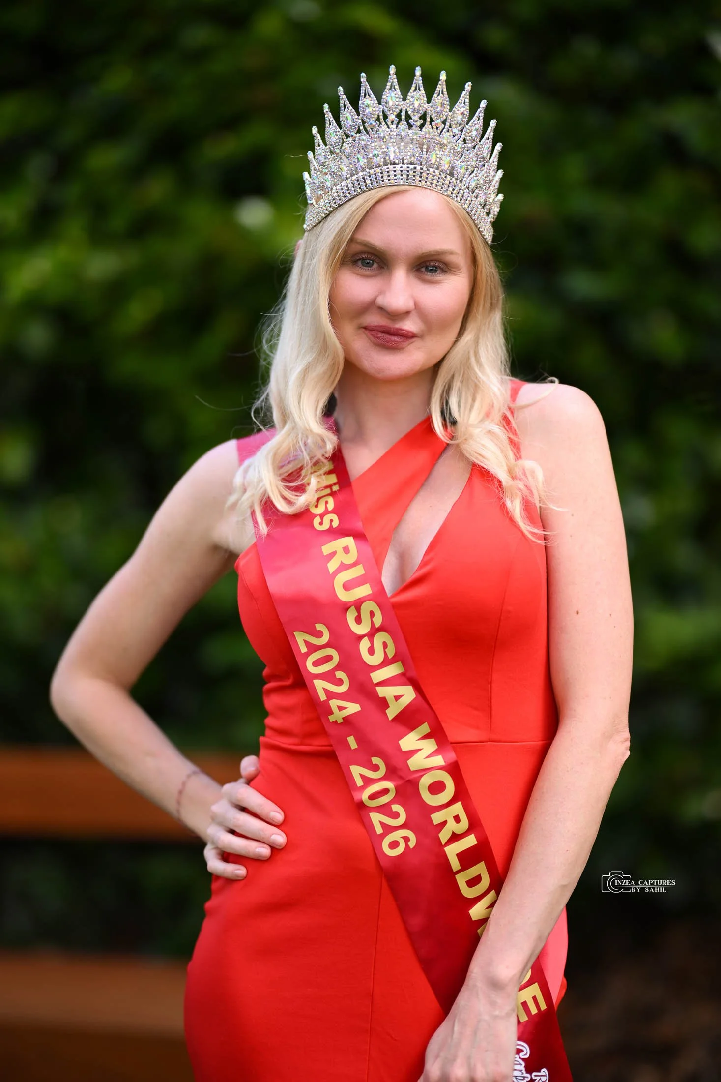 A woman with blonde hair wearing a crown and a red dress, with a sash that reads 'Miss Russia 2024 - 2026', standing outdoors with greenery in the background.