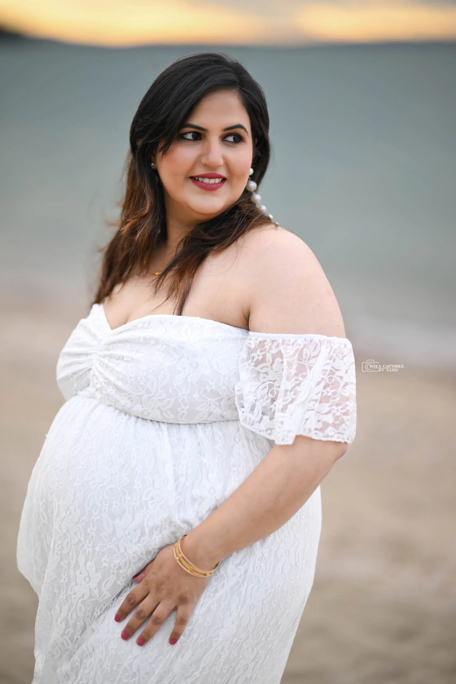 A pregnant woman in a white off-shoulder lace dress standing on a beach at sunset, smiling and looking to the side.