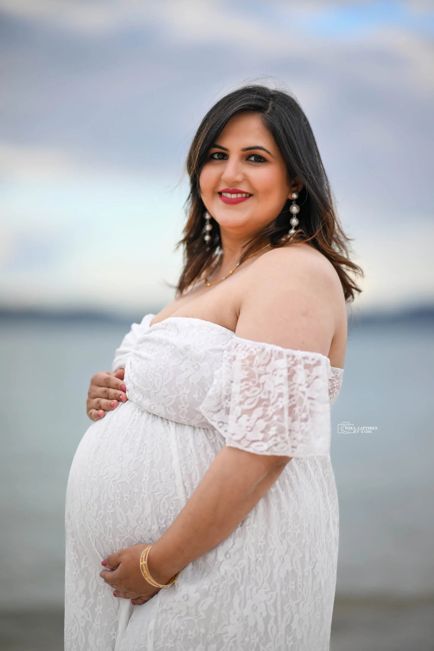 Pregnant woman in a white lace off-shoulder dress standing by the water, smiling, with dark hair, pearl earrings, and gold jewelry.