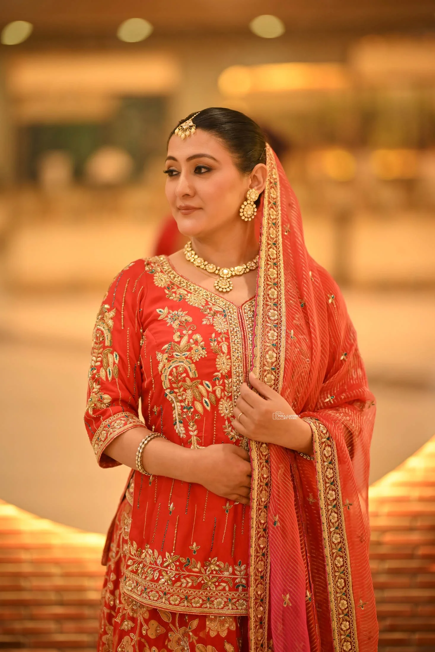 A woman dressed in a traditional red Indian outfit with gold and floral embroidery, accessorized with gold jewelry, standing indoors with a blurred background.
