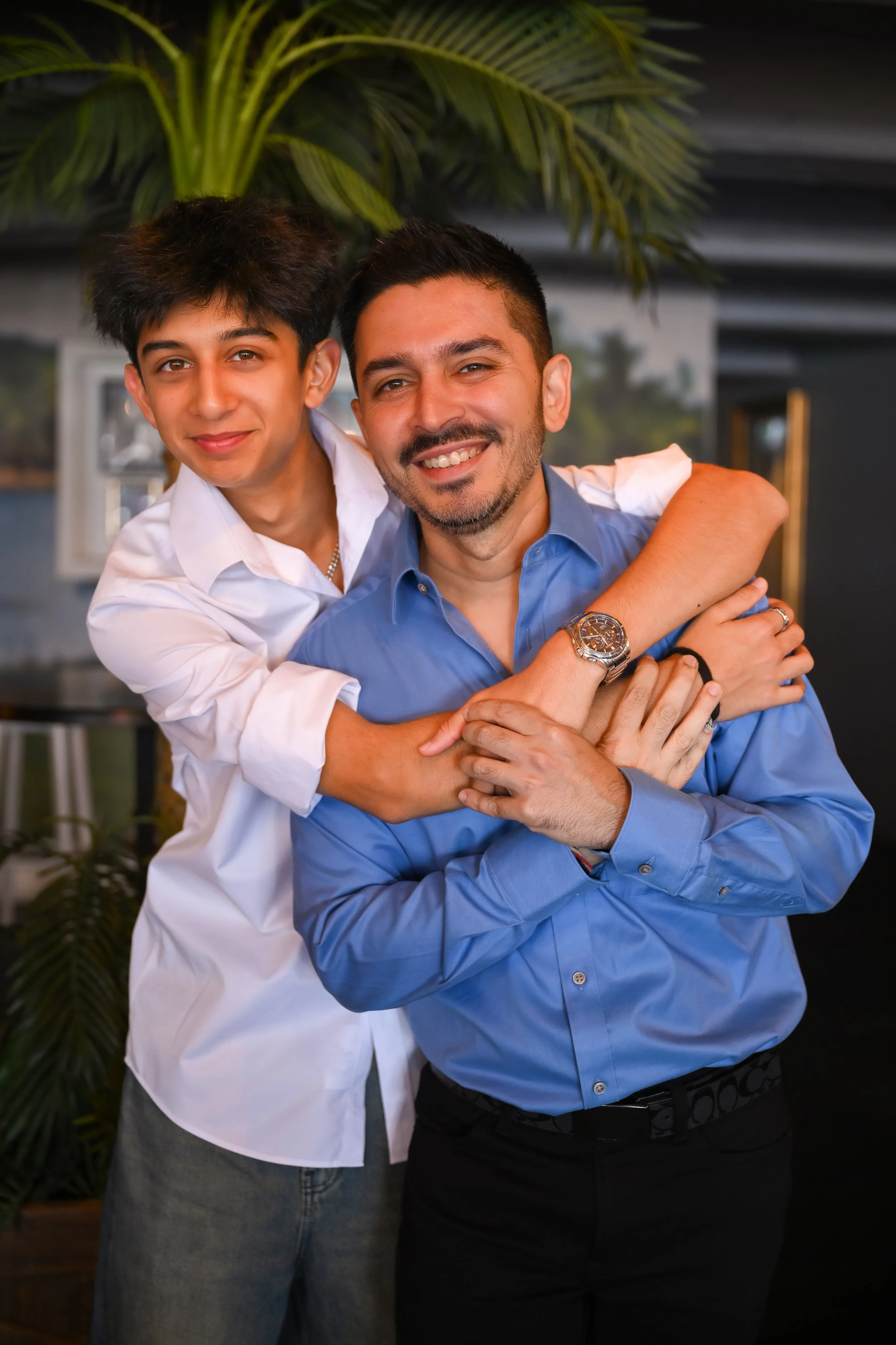 A young man and an older man, possibly father and son, smiling and embracing each other inside a room with tropical plants in the background.