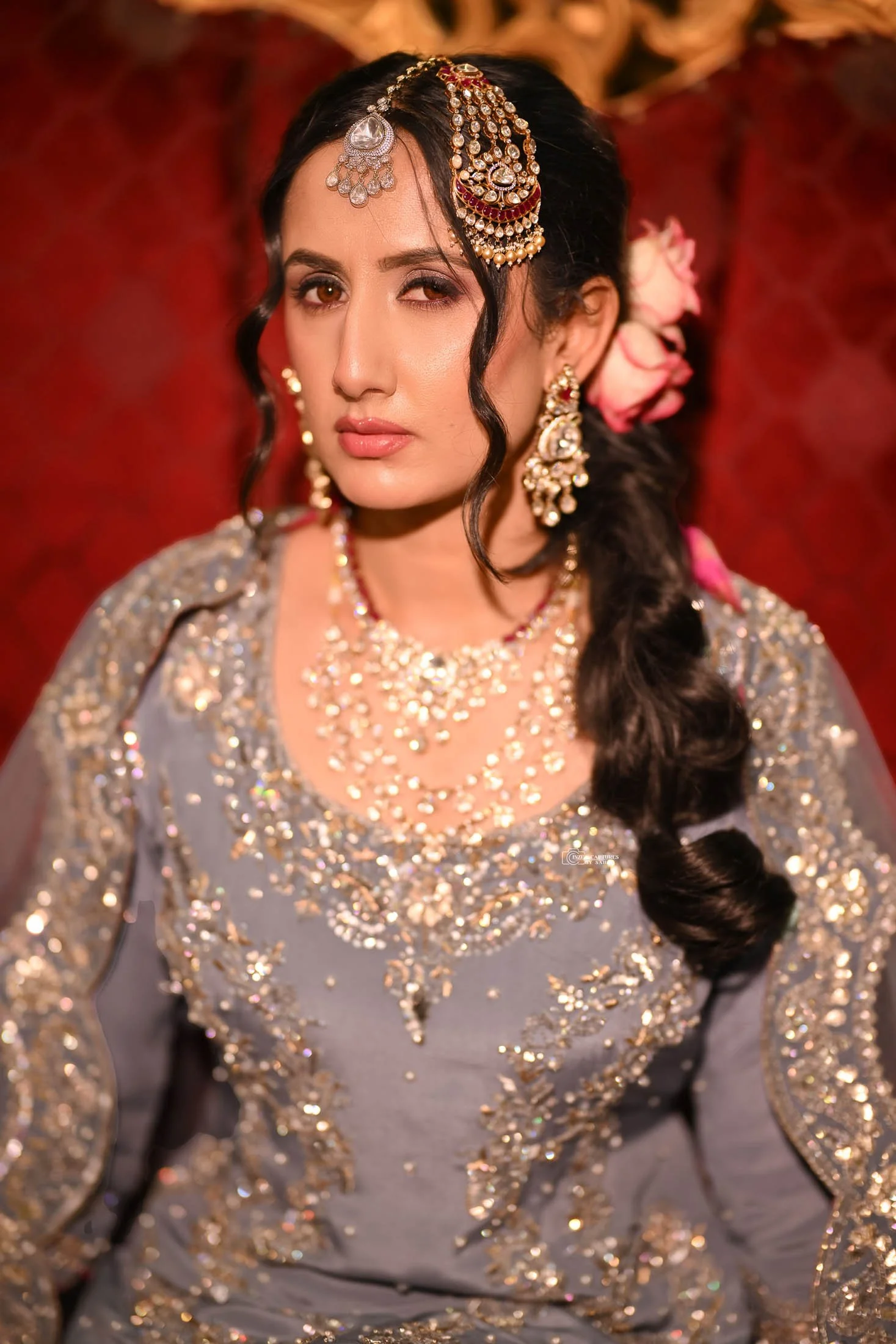 A woman dressed in traditional Indian wedding attire, wearing heavy jewelry including earrings, necklaces, and a decorated headpiece. She has long dark hair styled in a braid with pink flowers and a slightly tilted head, with a confident expression a
