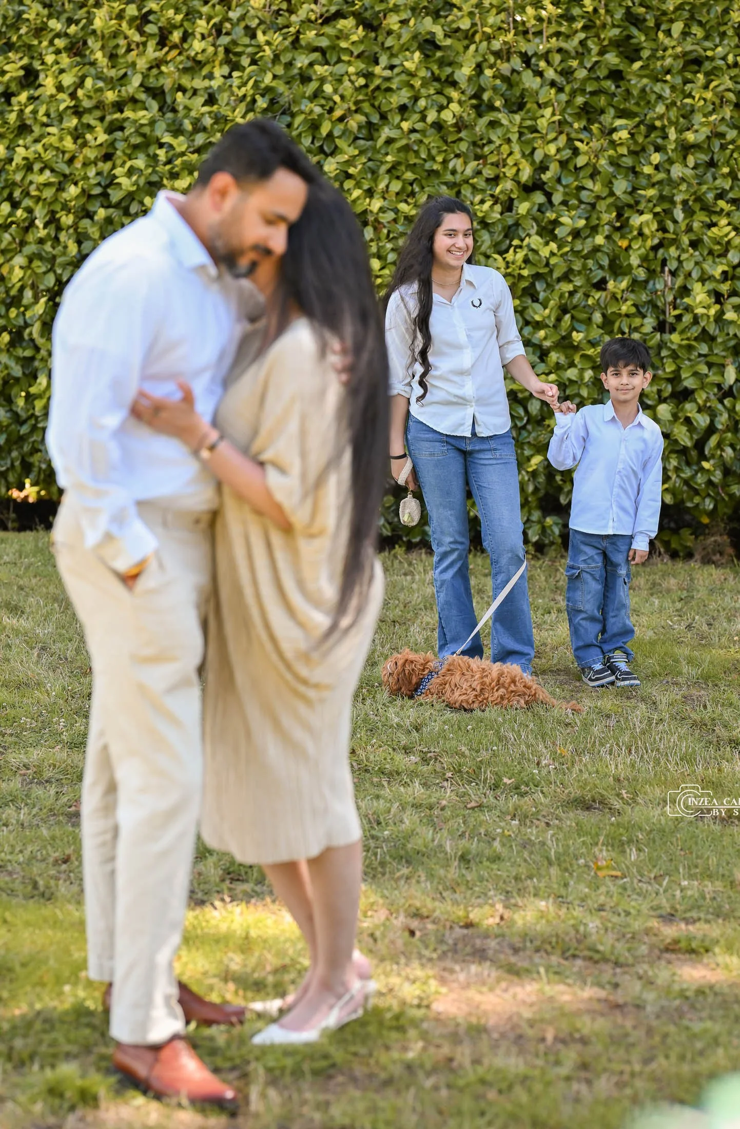 A family outdoors, with a man and woman embracing in the foreground, and a young woman and boy holding hands with a small brown dog lying on the grass in the background, in front of a green hedge.