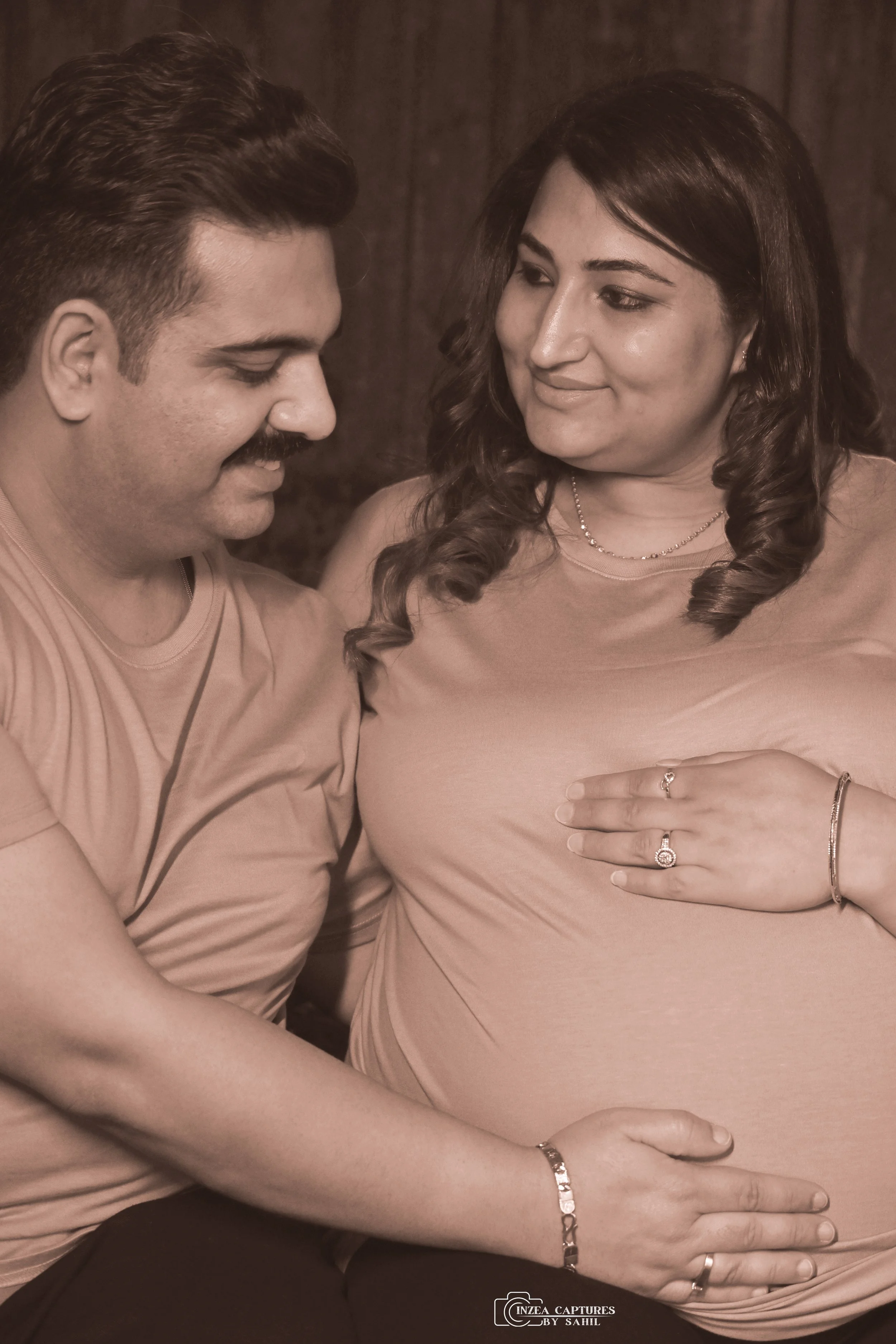 A happy couple sharing an intimate moment, with the woman showing her pregnant belly, both smiling and looking at each other, in a warmly lit setting.