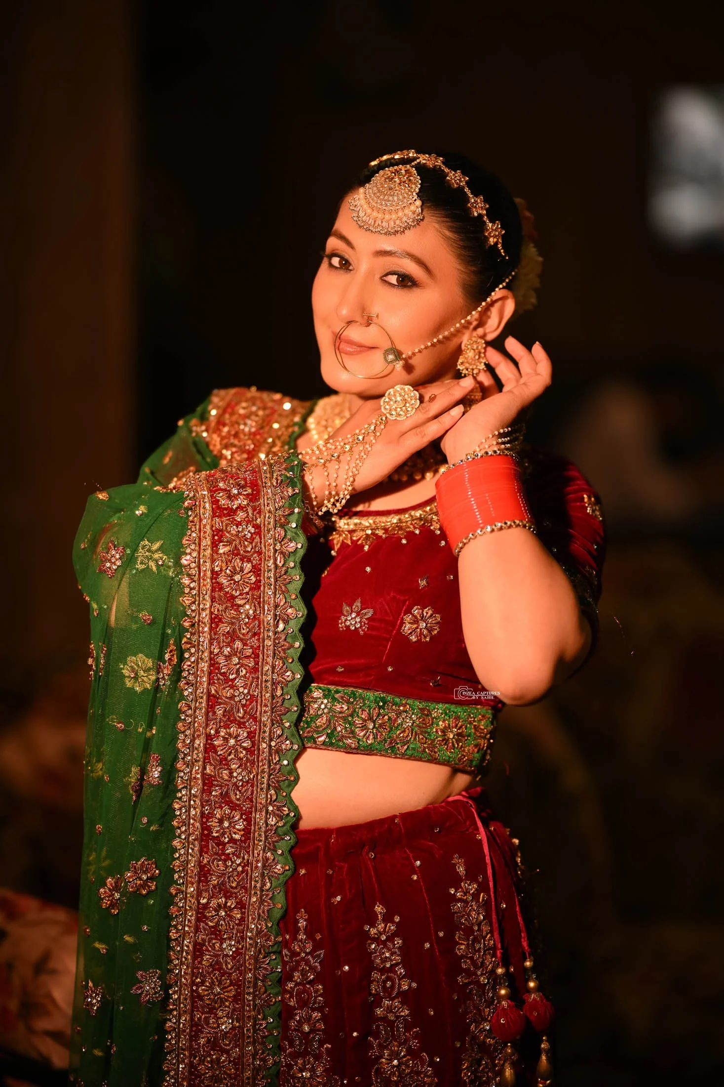 A woman dressed in traditional Indian bridal attire, wearing a red and green embroidered lehenga with gold jewelry and accessories, smiling and posing gracefully.