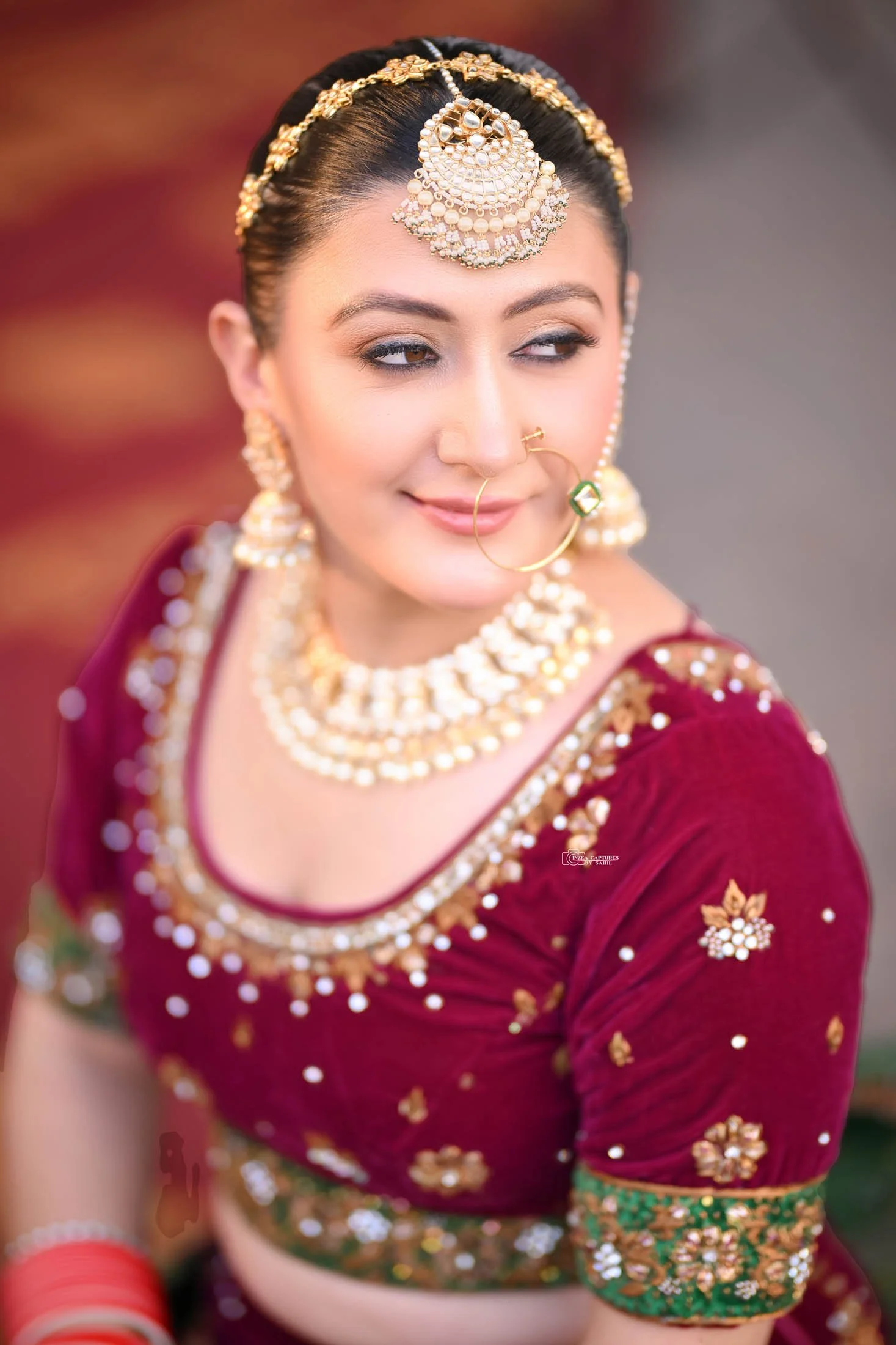 A woman dressed in traditional Indian bridal attire with intricate gold and pearl jewelry, a maroon and gold embroidered blouse, and a matching veil, smiling softly and looking to the side.