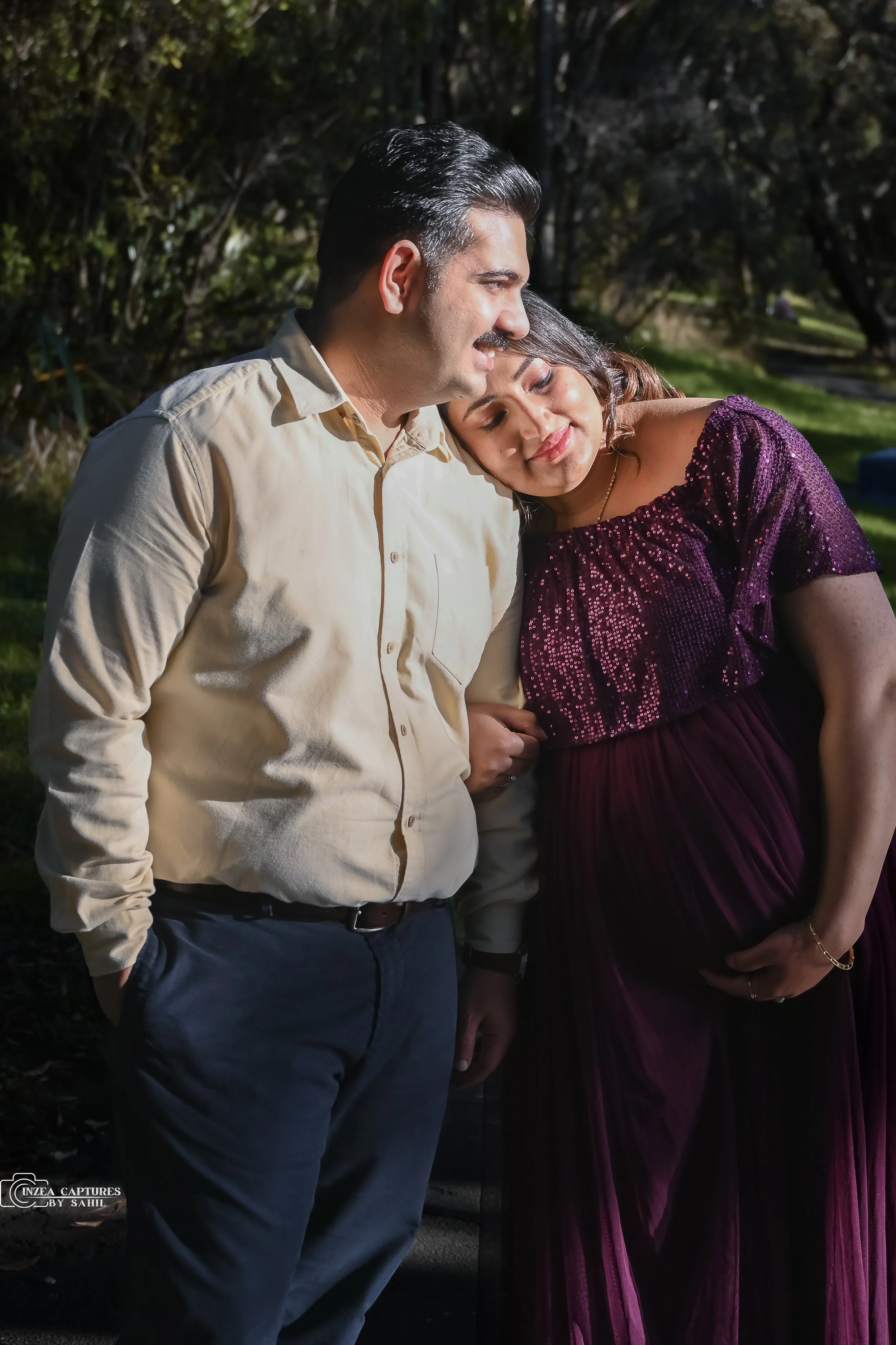 A couple standing close together outdoors, with the woman resting her head on the man's shoulder and smiling, during the daytime.