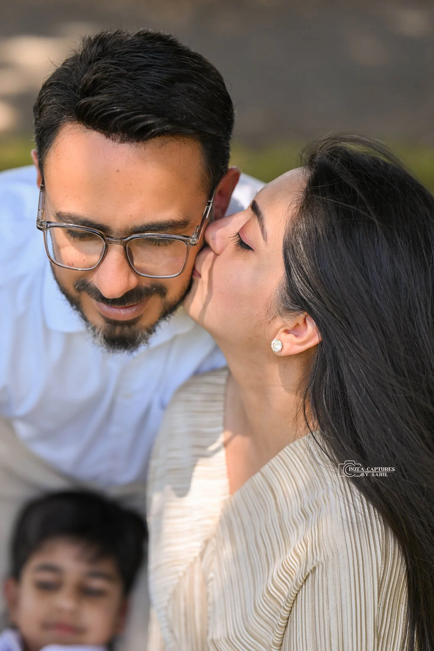 A woman kissing a man on the cheek outdoors, with a young boy in the foreground.