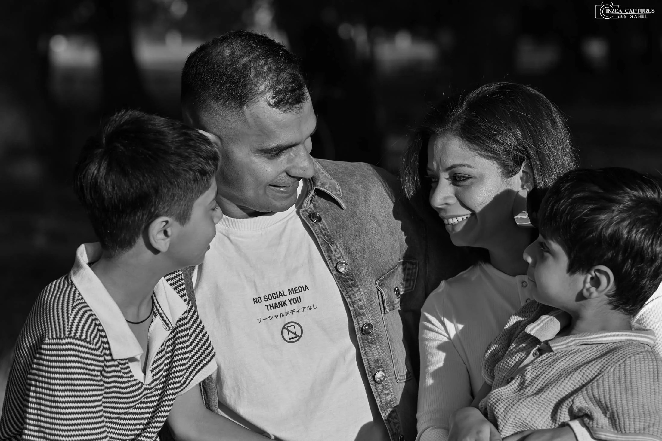 A family of four, including a man, a woman, and two boys, sharing a happy moment outdoors. The man is wearing a T-shirt with the text 'NO SOCIAL MEDIA THANK YOU' and Japanese characters, and a denim jacket. The woman has short hair and is smiling at 