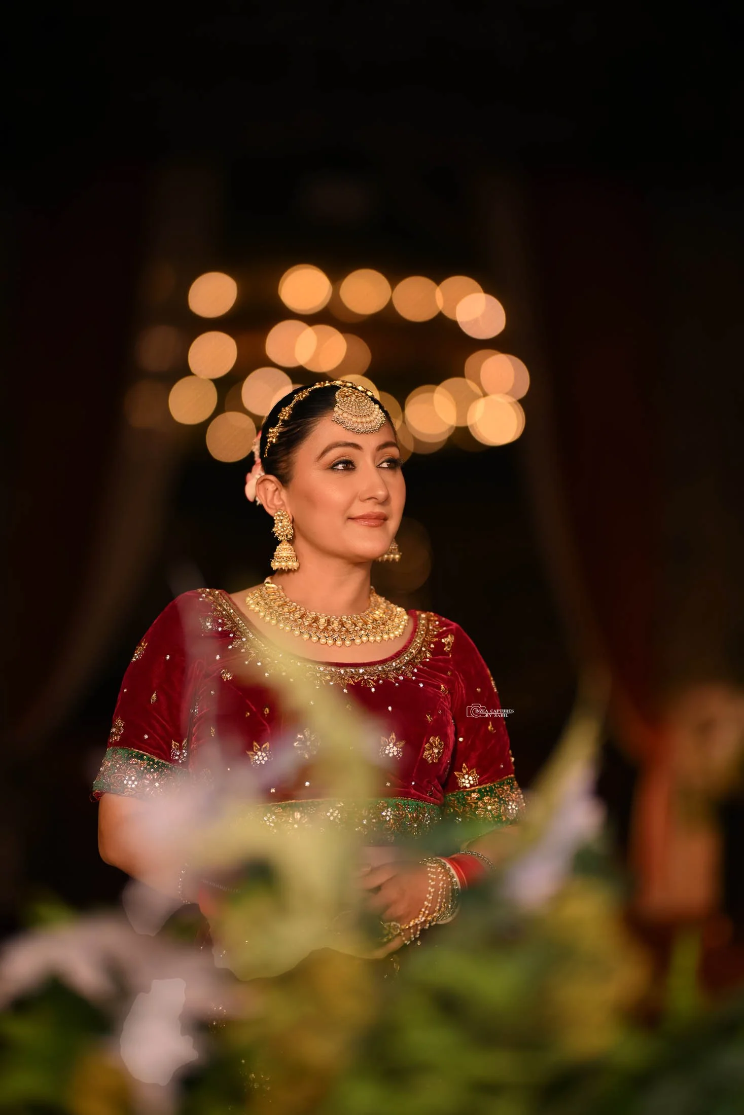 A woman dressed in traditional Indian attire with jewelry, standing in a dimly lit setting with blurred warm string lights in the background.
